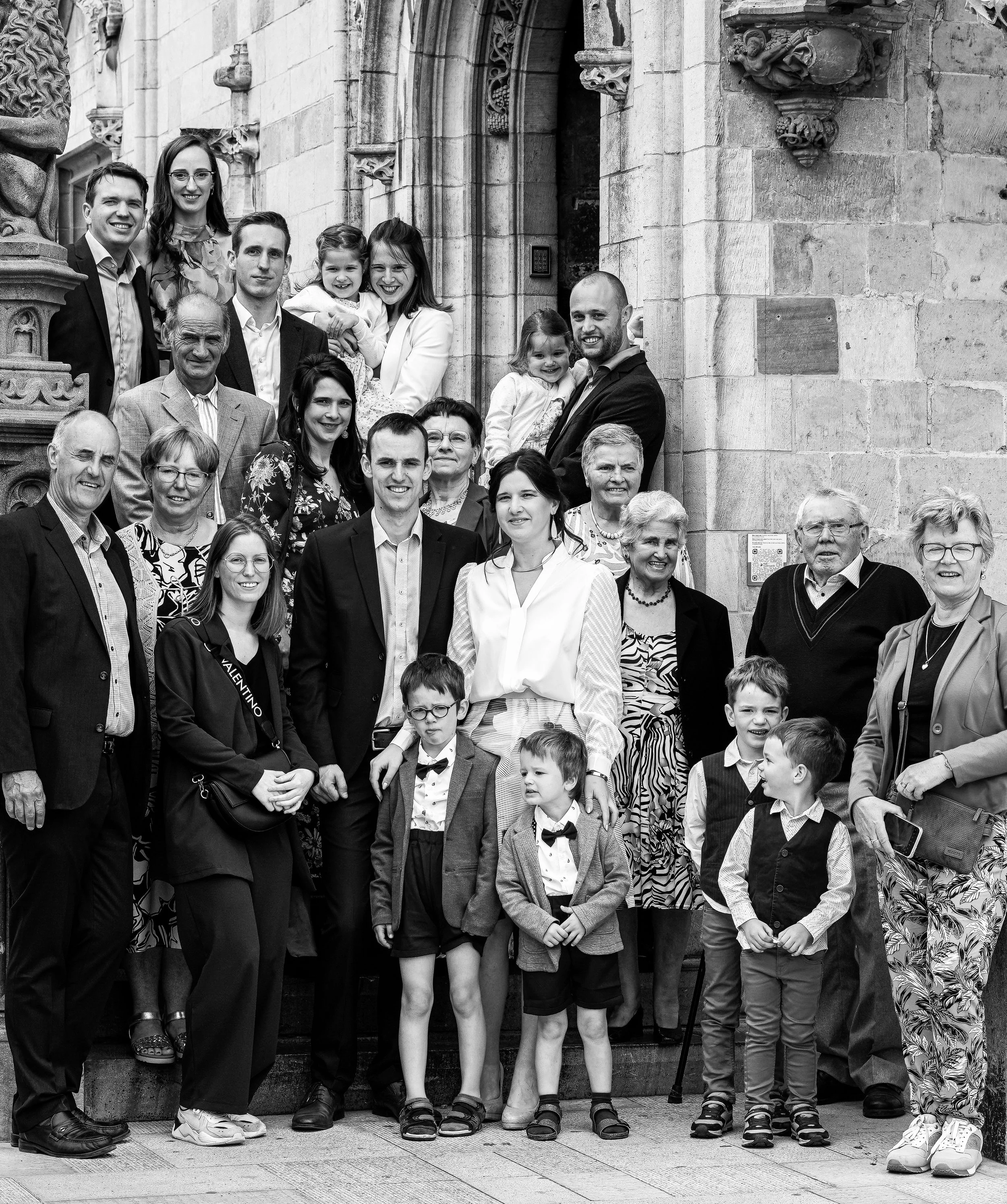 Groep mensen poseert voor een historische stenen achtergrond, inclusief volwassenen, kinderen en ouderen, in zwart-wijs.