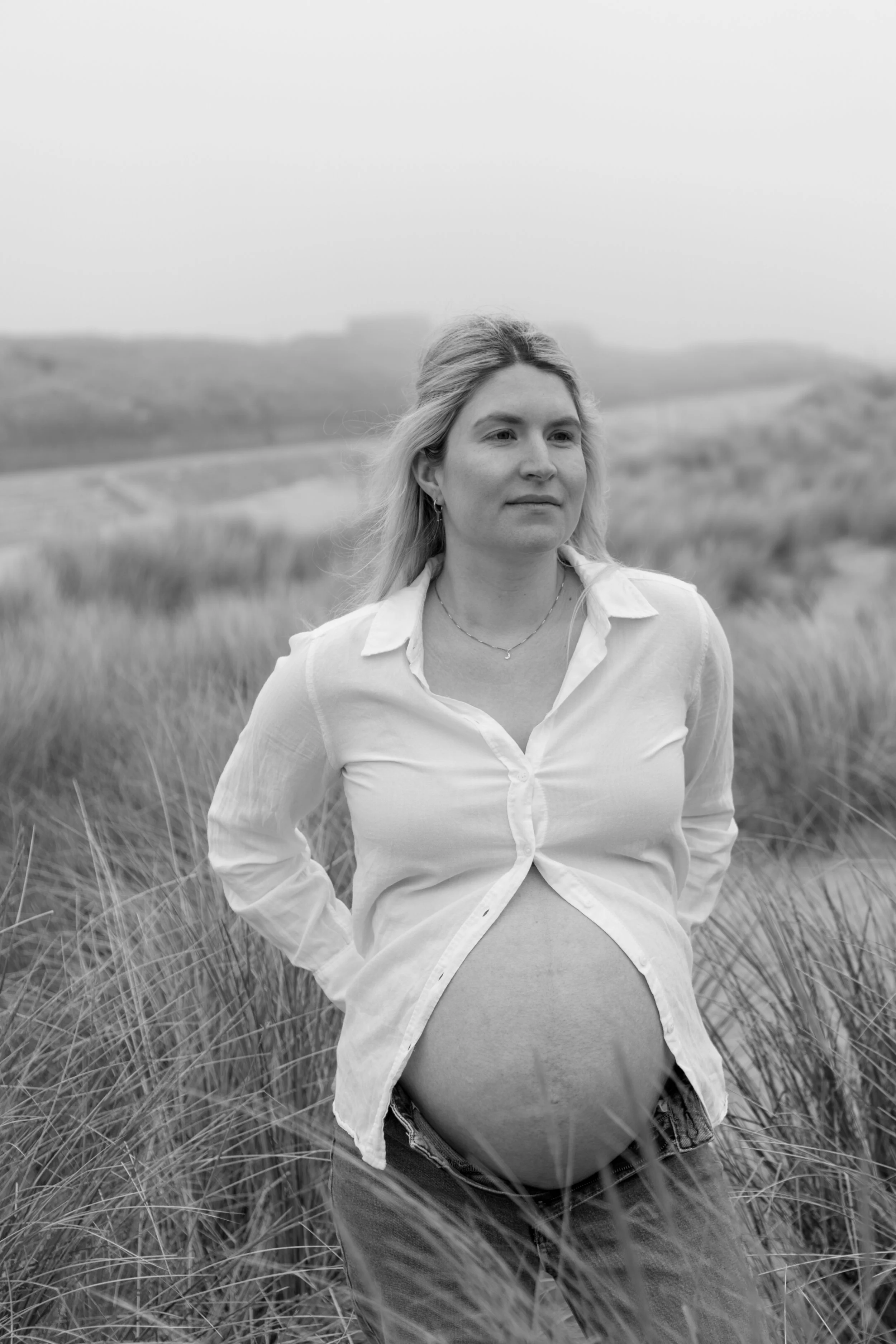 Zwart-wit foto van een zwangere vrouw die in een grasland staat, met een mistige achtergrond en een heuvel op de horizon.