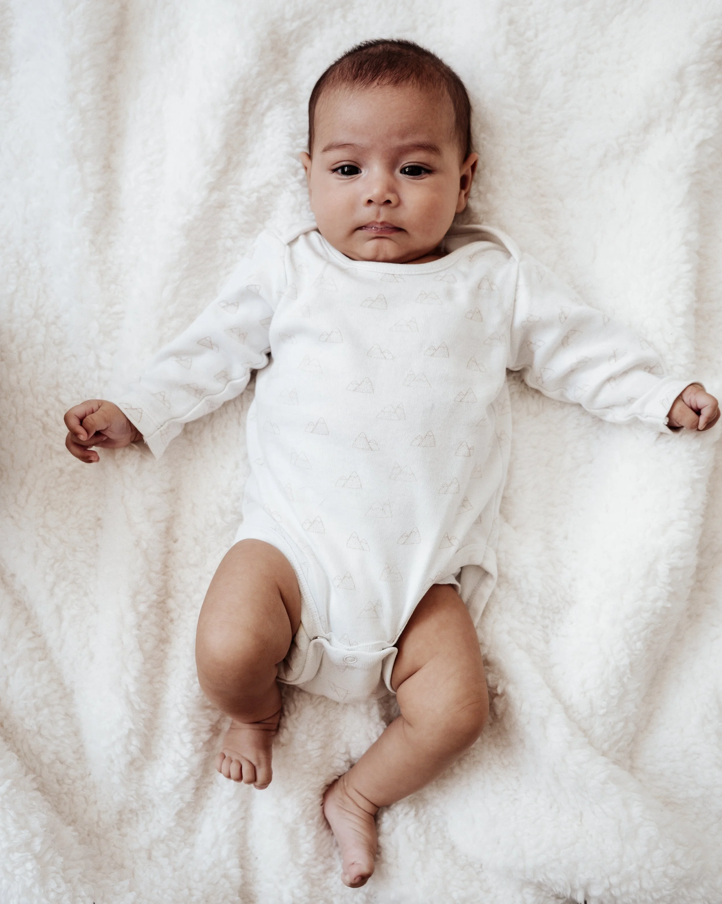 Een baby ligt op een zachte, crèmekleurige deken, draagt een witromp met een subtiel patroon en kijkt naar de camera.