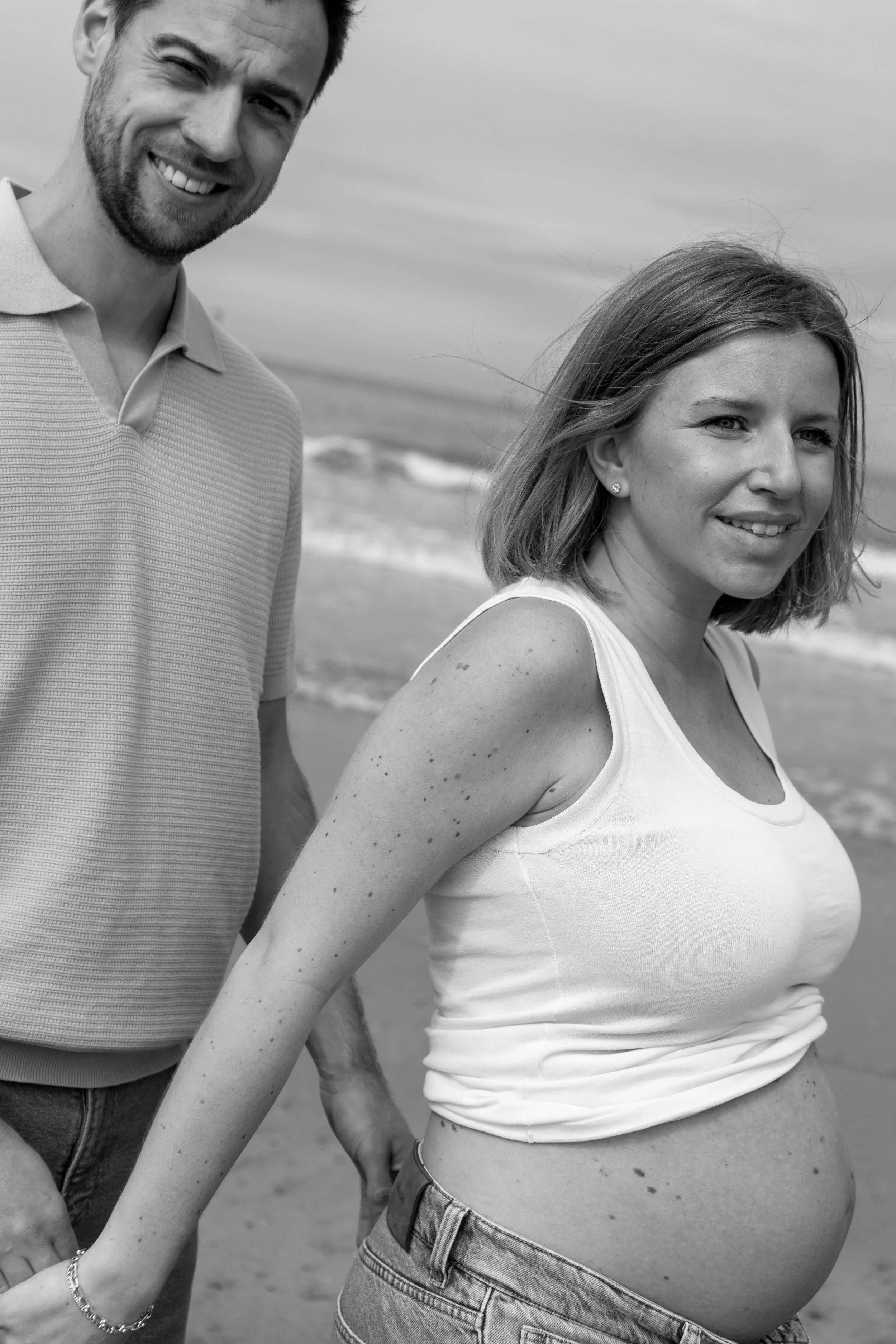 Een man en een zwangere vrouw staan op het strand, lachend en poseren voor de foto.