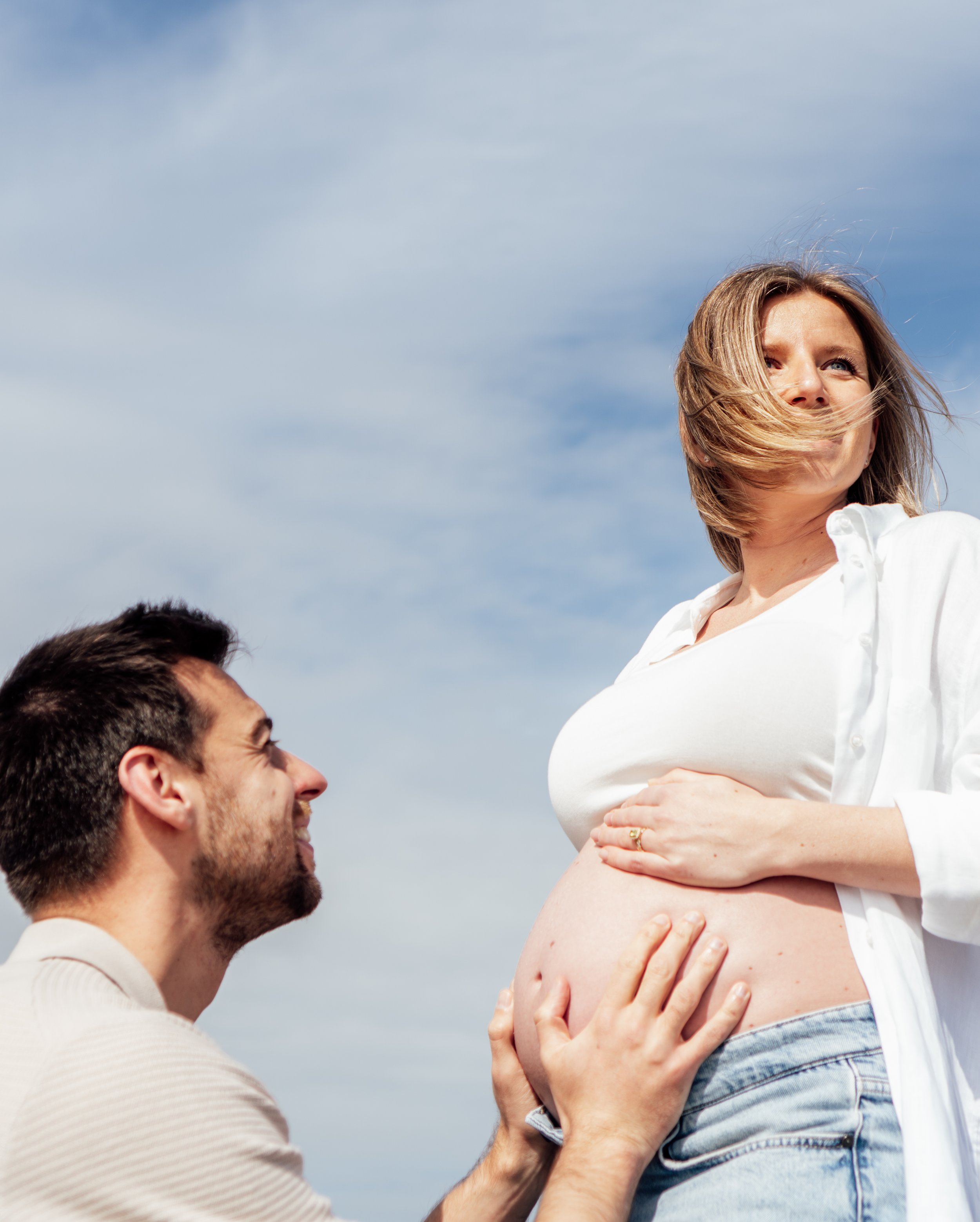 Gelukkige man knielt voor zwangere vrouw die haar buik vasthoudt en lachend kijkt, met een blauwe lucht op de achtergrond.