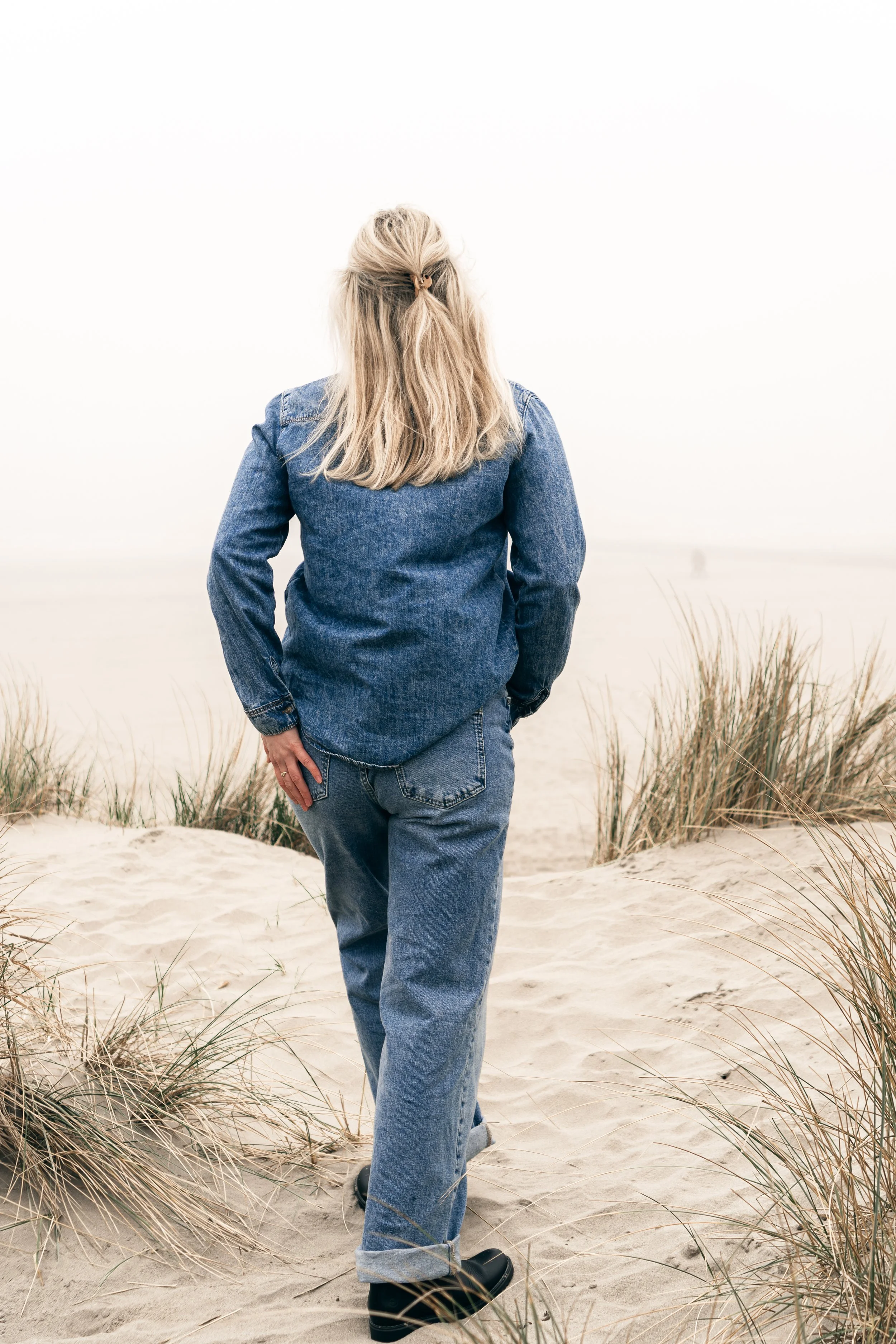 Persoon in denim jas en jeans wandelt door zand op een strand, met droog gras en een bewolkte sky op de achtergrond.