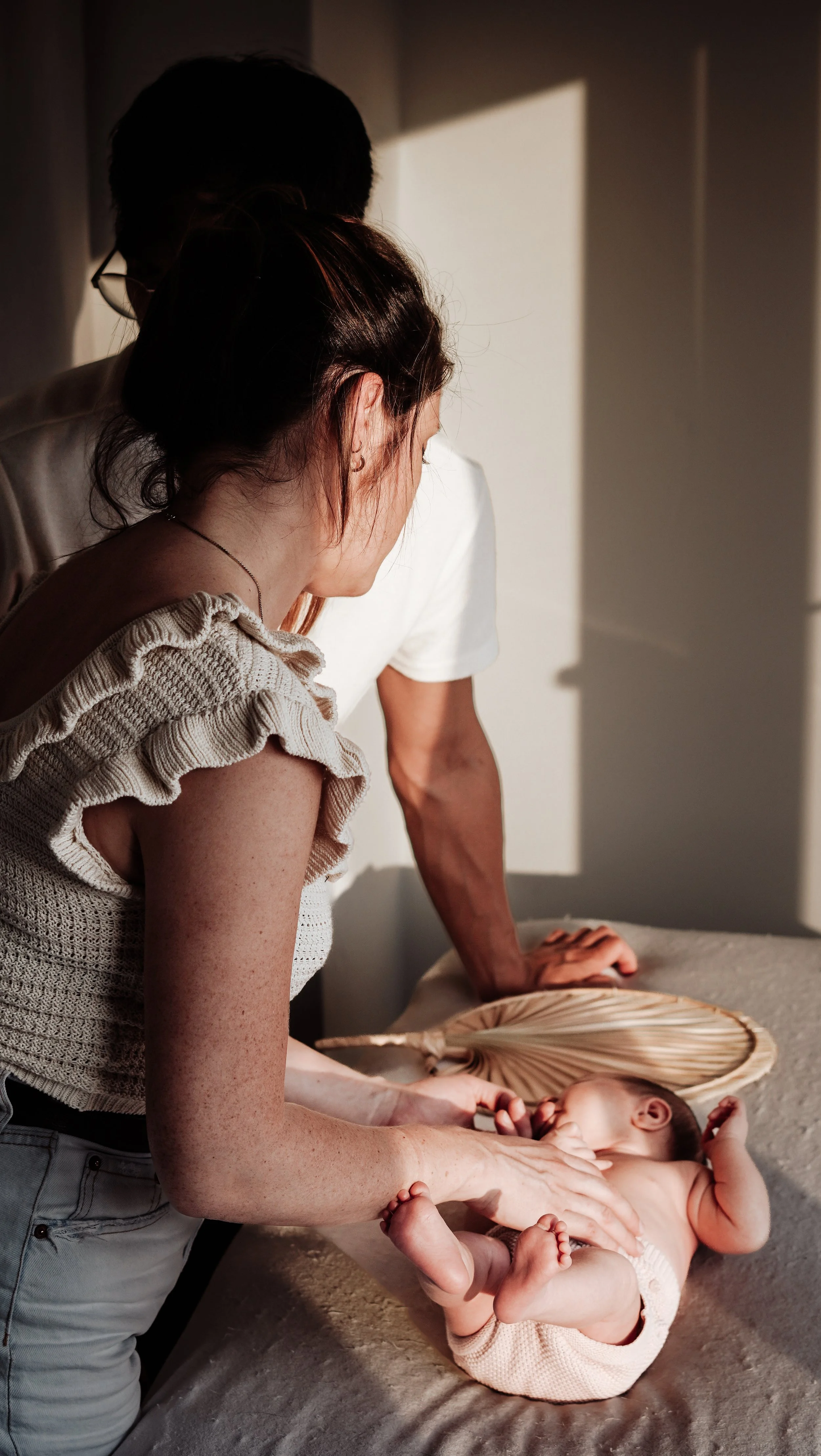Een baby wordt verzorgd door twee volwassenen op een bed, met zonlicht dat door een raam naar binnen valt.