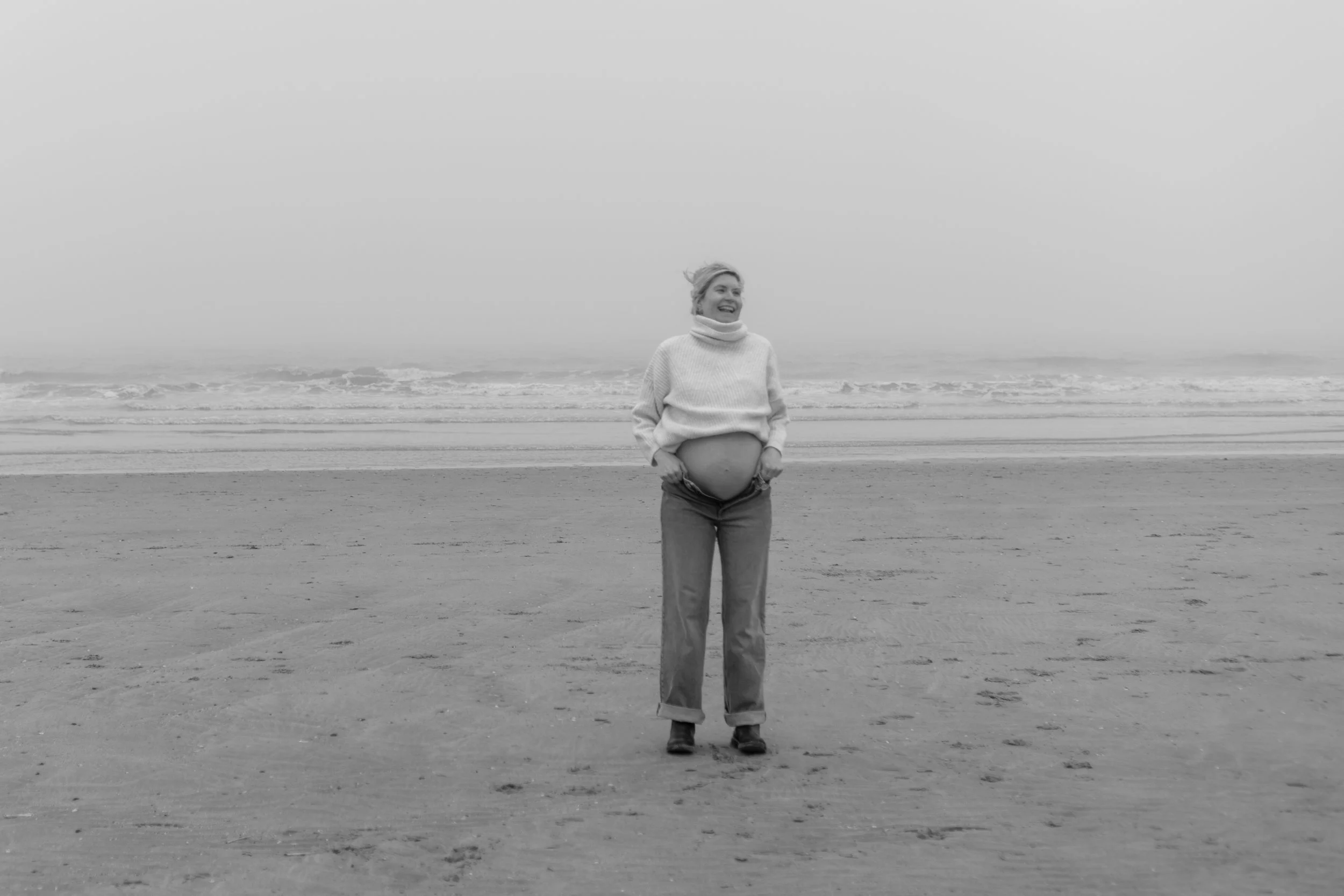 Een zwart-wit foto van een vrouw op het strand met een grote buik, ze glimlacht en draagt een witte trui en jeans, terwijl de zee op de achtergrond te zien is.