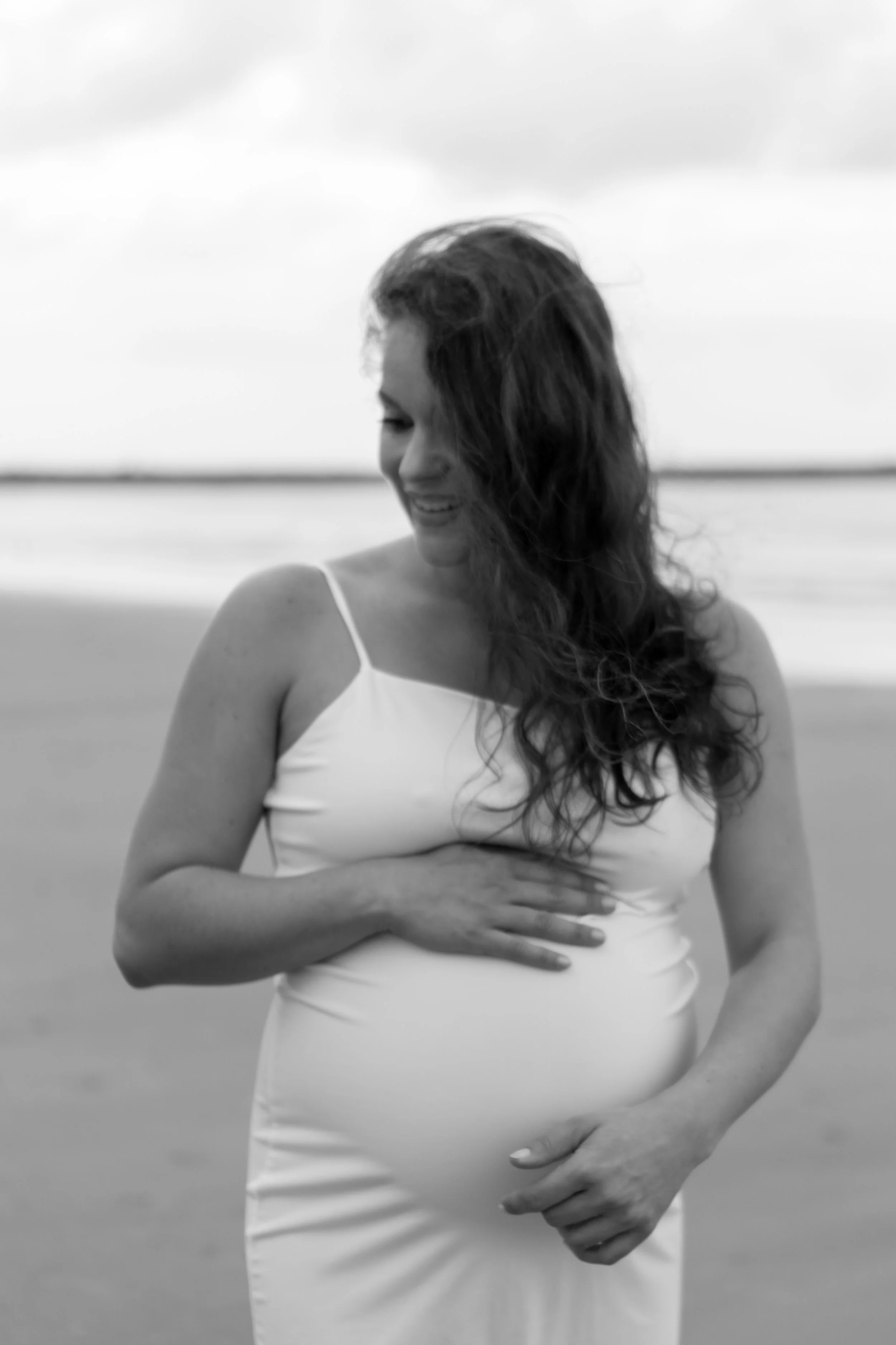 Zwangere vrouw in witte jurk die haar buik vasthoudt, op het strand met de zee op de achtergrond, in zwart-win