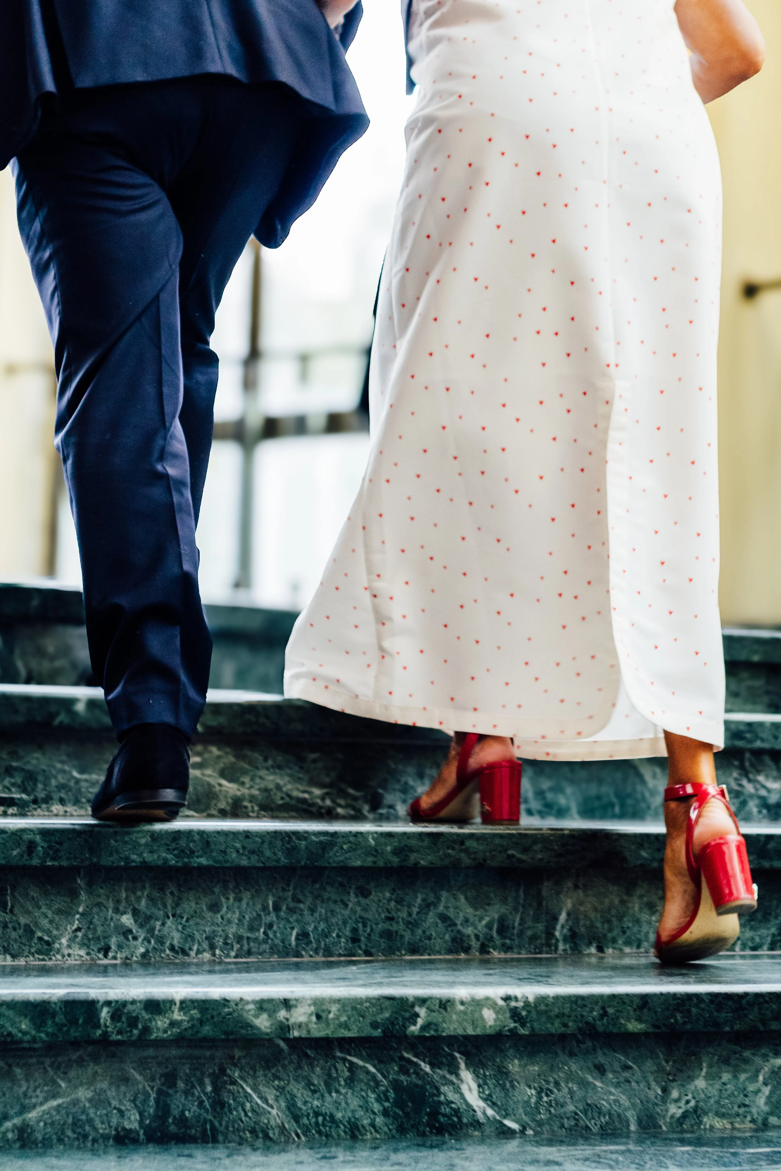 De afbeelding toont de onderkant van twee mensen die de trap op lopen. Eén persoon draagt een witte rok met rode stippen en rode high heels, de andere persoon draagt een blauwe pak en zwarte schoenen.