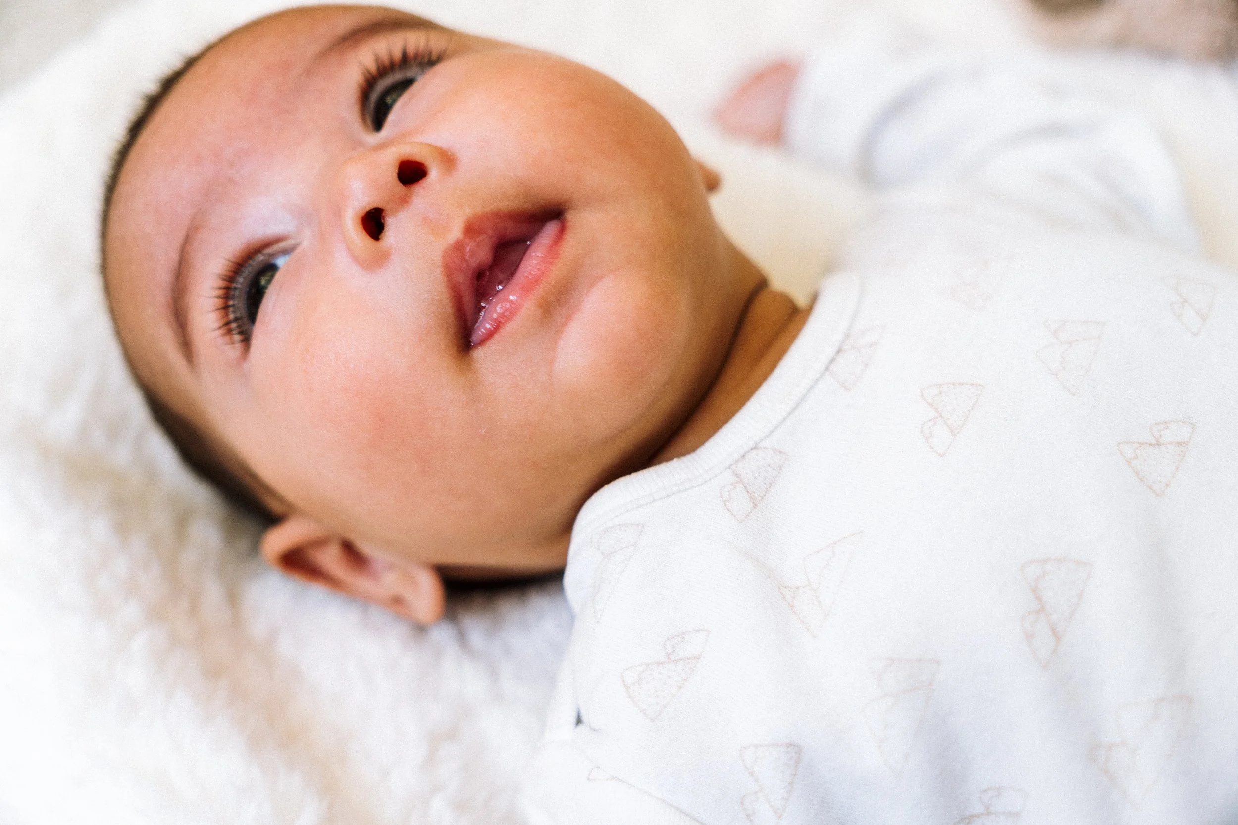 Close-up van een lachende baby met grote ogen, liggend op een witte, zachte ondergrond, gekleed in een wit pakket met kleine hartjespatronen.