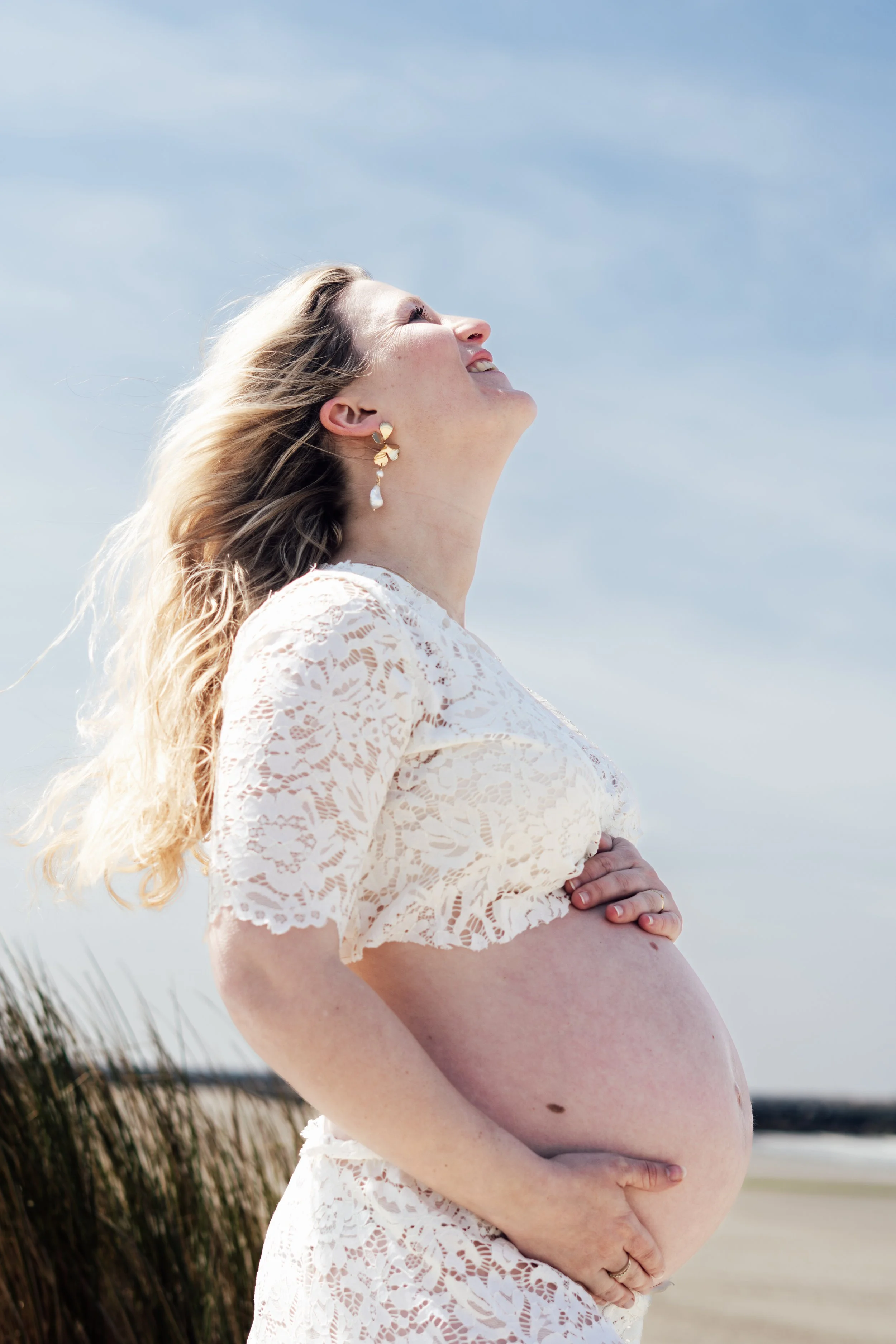 Zwarte en witte foto van een vrouw met een chaotische hairstyle, een zwarte jas, een zwarte lingerie en een ketting met een bloemvormige hanger. Ze zit op een bank en kijkt naar rechts.
