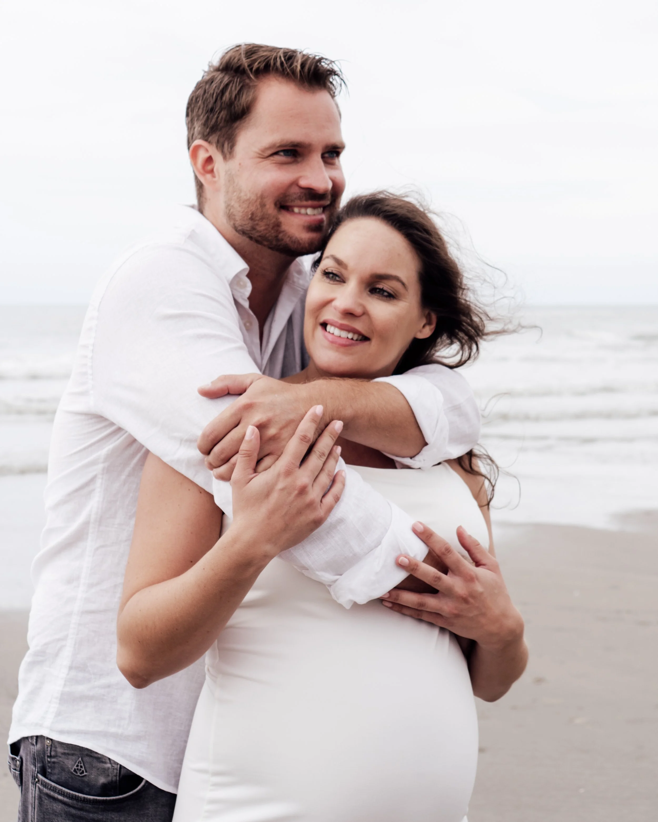 Een zwangere vrouw en een man omhelzen elkaar aan het strand.