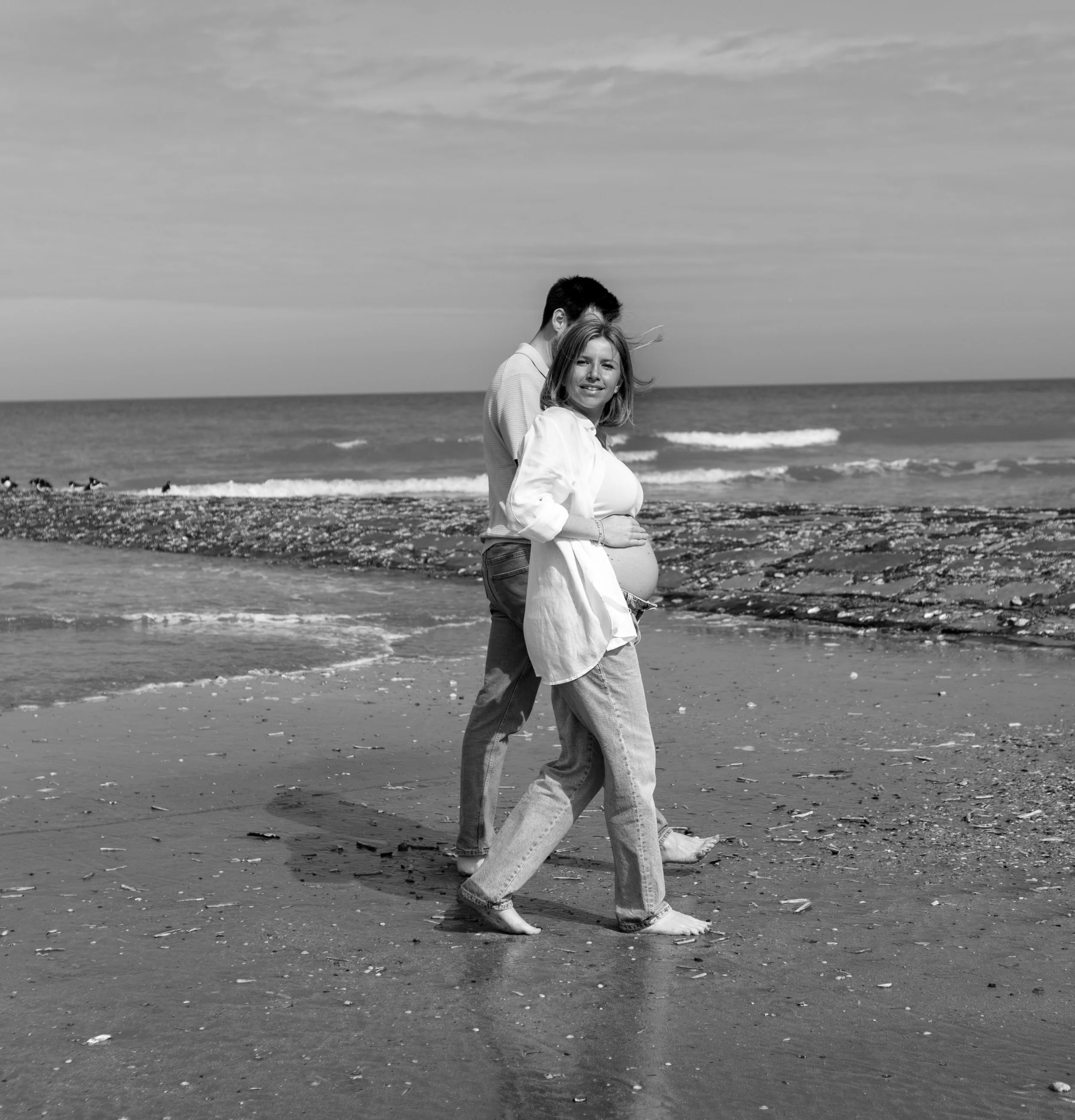 Zwart-wit foto van een zwangere vrouw en haar partner die samen op het strand lopen, met de oceaan op de achtergrond.