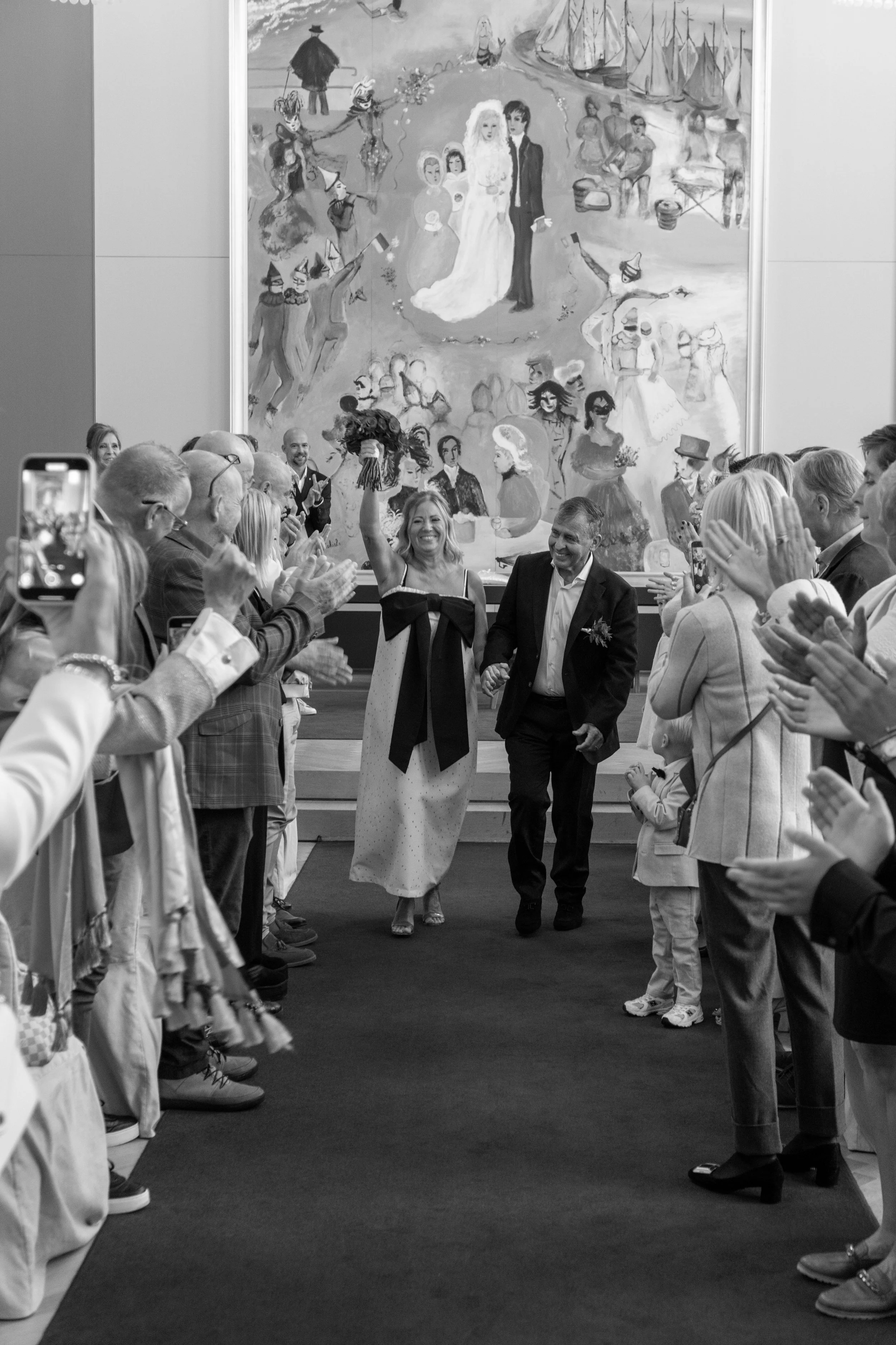 Mensen staan in een hal en applaudisseren naar een paar dat in het midden onder een grote schildering of muurschildering staat. De mensen dragen formele en feestelijke kleding, en de sfeer lijkt feestelijk of bijzonder.