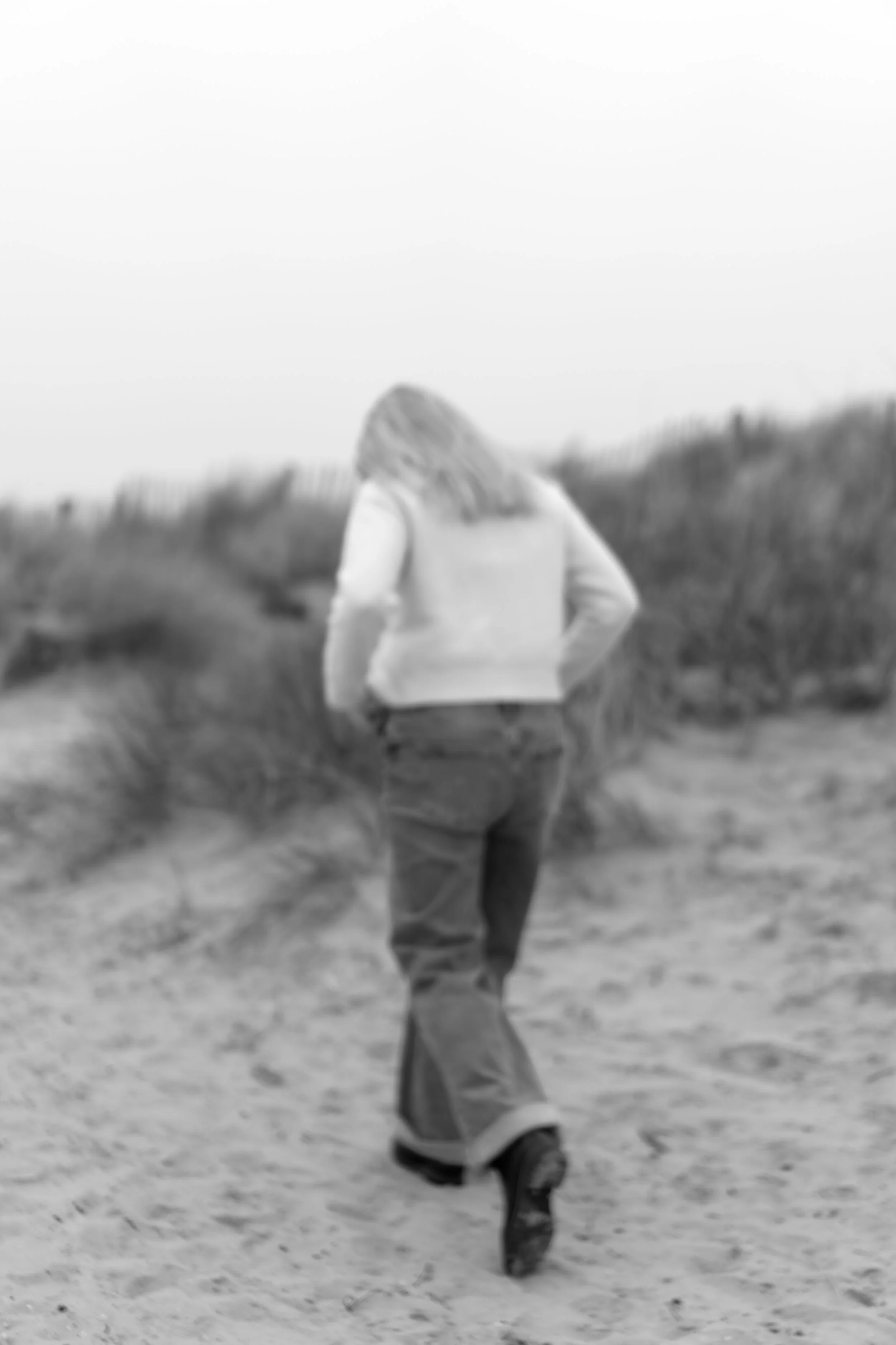 Een persoon die op een skateboarding staat op een strand of duinen, met lange haar en een jas.