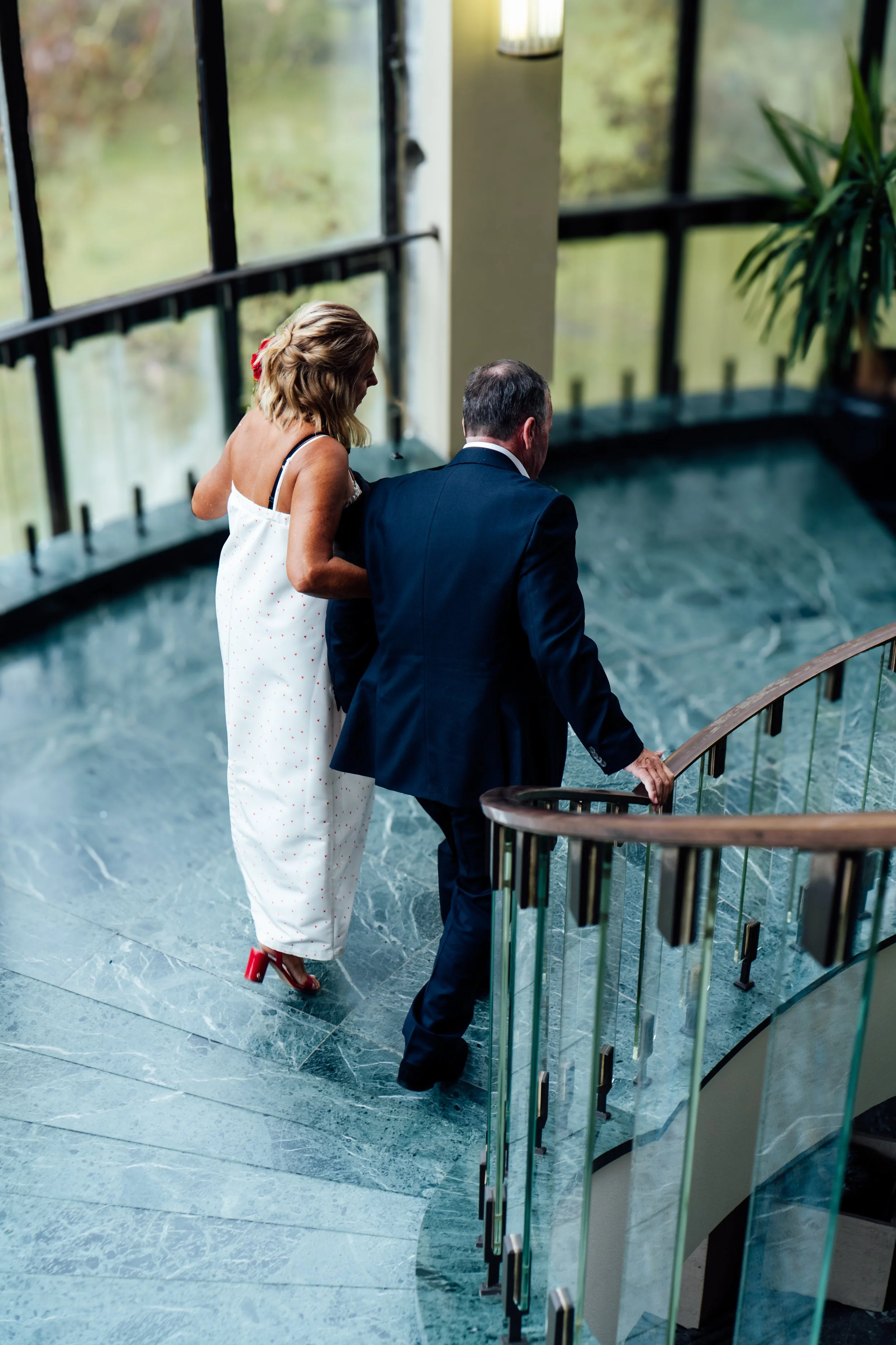 Een vrouw in een witte jurk met rode schoenen en een man in een zwart pak lopen samen op een wenteltrap met een glazen reling, binnen, met grote ramen en een plant op de achtergrond.