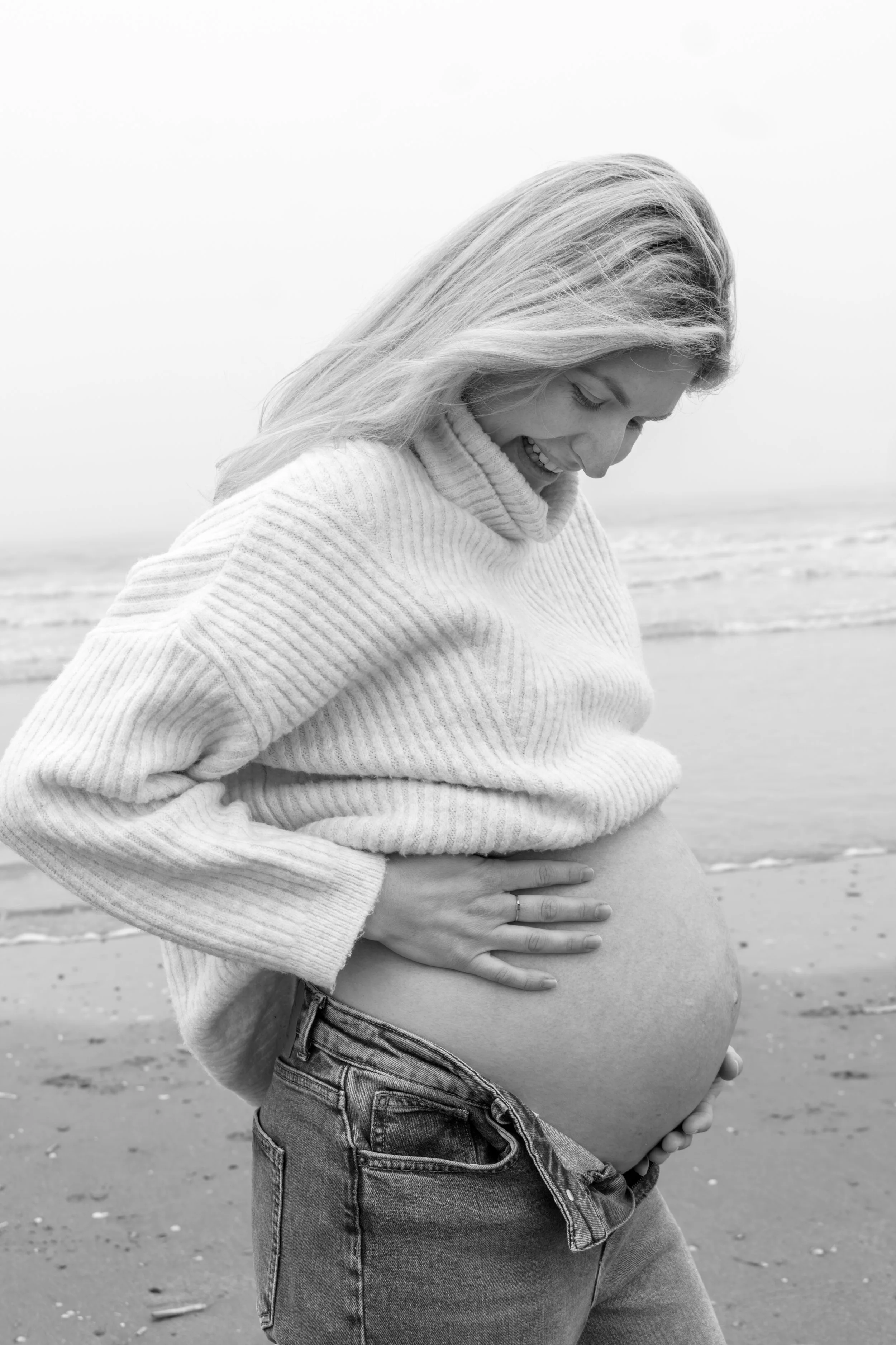 Een zwangere vrouw op het strand, glimlachend en met haar hand op haar buik.