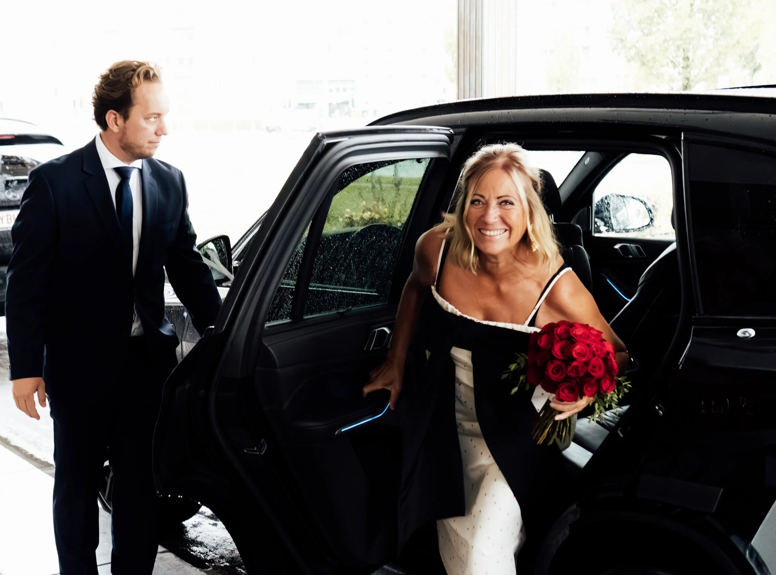 Lachende vrouw met bloemen in de auto, man in pak staat erbij, regenachtig weer buiten.