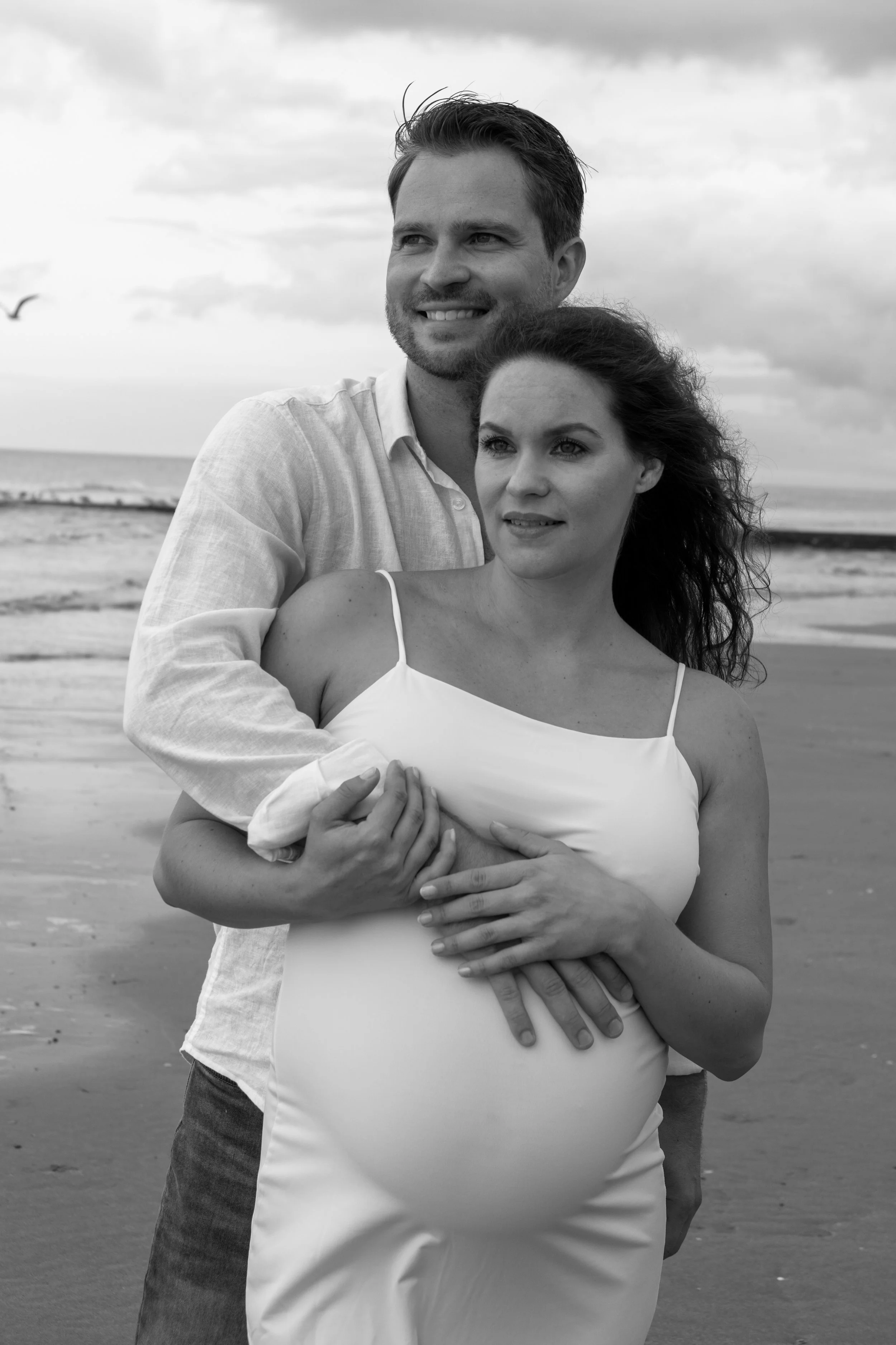 Een paar poseert samen op het strand, de man staat achter de vrouw en heeft zijn armen om haar heen, beiden kijken vriendelijk en blij.