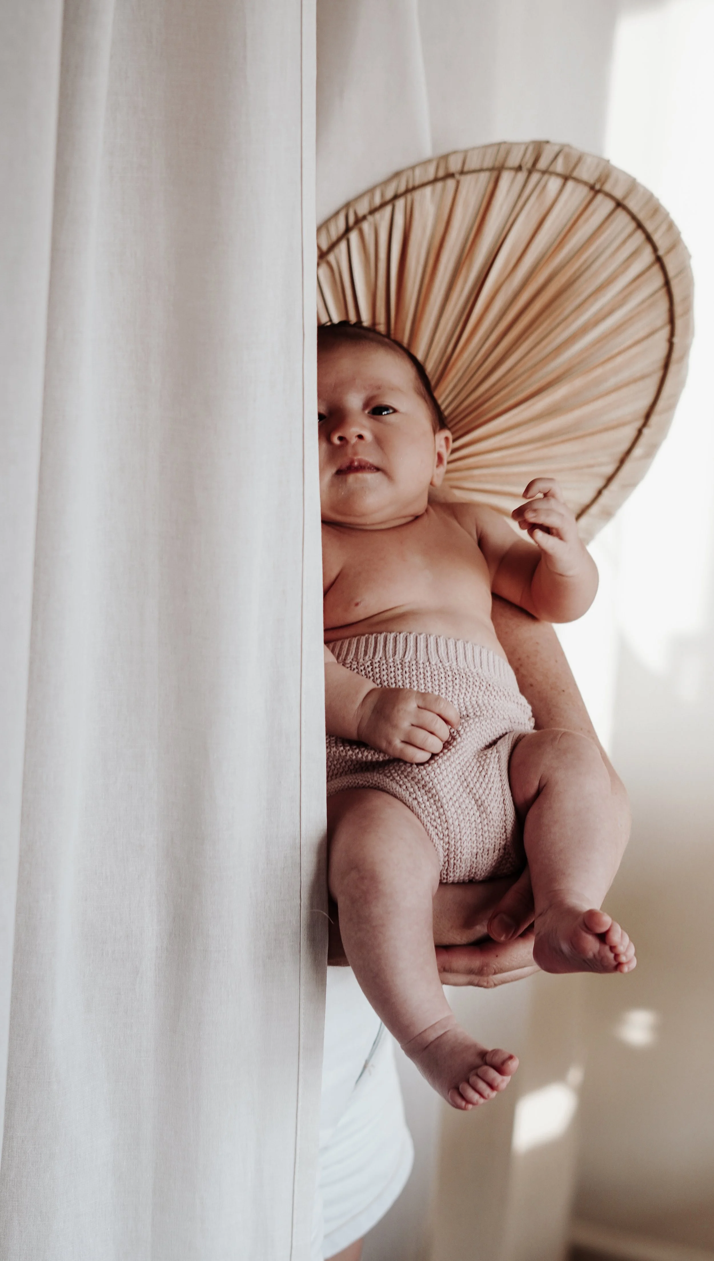 Kleintje met een hoofdband die eruit ziet als een grote paddestoel, zittend op een volwassen arm, half zichtbare persoon achter een witte gordijn.