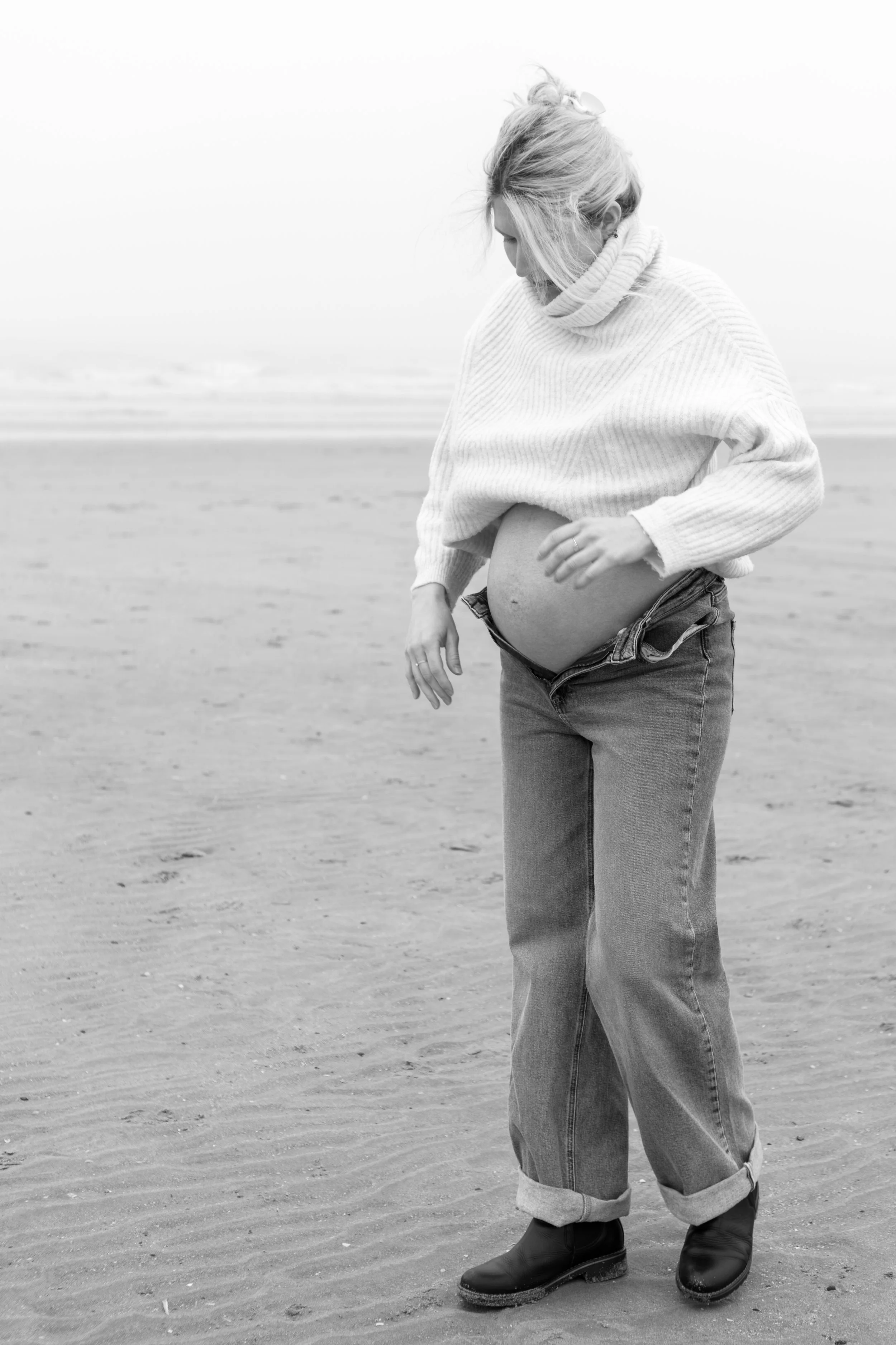 Vrouw op het strand draagt een dikke, witte gebreide trui, jeans en stevige zwarte laarzen, met haar buik zichtbaar, alsof ze zwanger is.