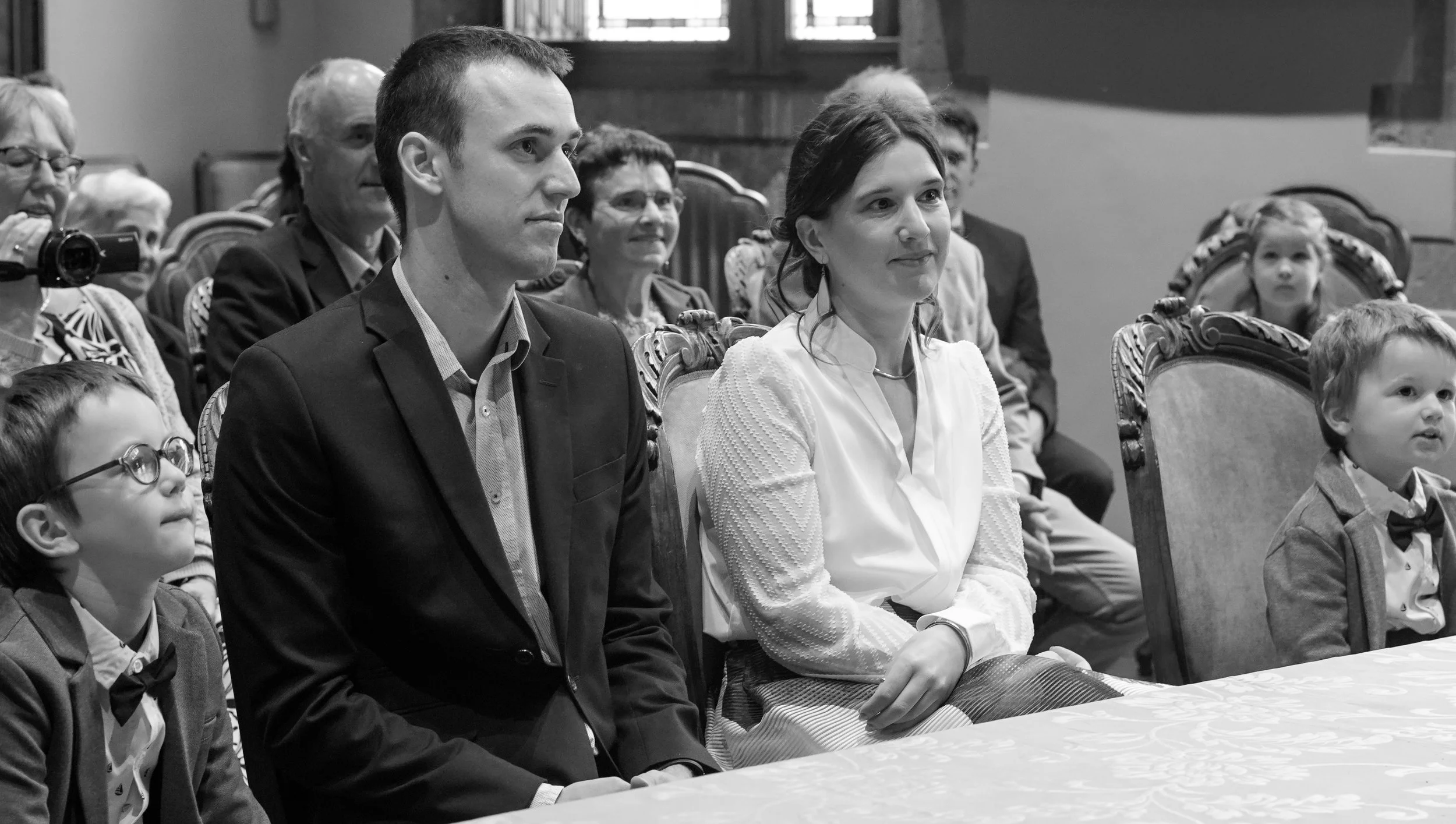 Een groep mensen, inclusief kinderen en volwassenen, zit attent en serieus aan een tafel in een binnenruimte, waarschijnlijk tijdens een bijeenkomst of evenement.