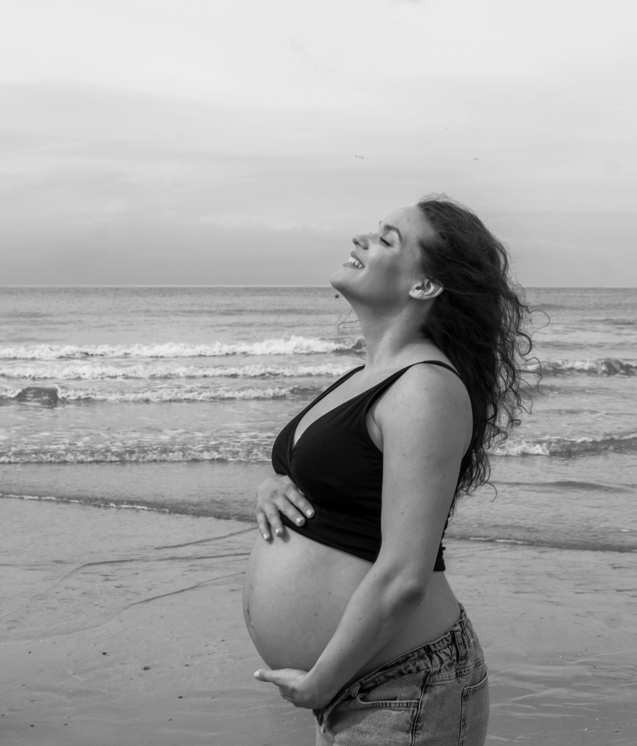 Zonovergoten vrouw met open ogen op een strand, lachend en in een zwarte top en spijkerbroeken, met haar handen op haar buik, terwijl ze de wind in haar haren voelt, met de zee en de lucht op de achtergrond.