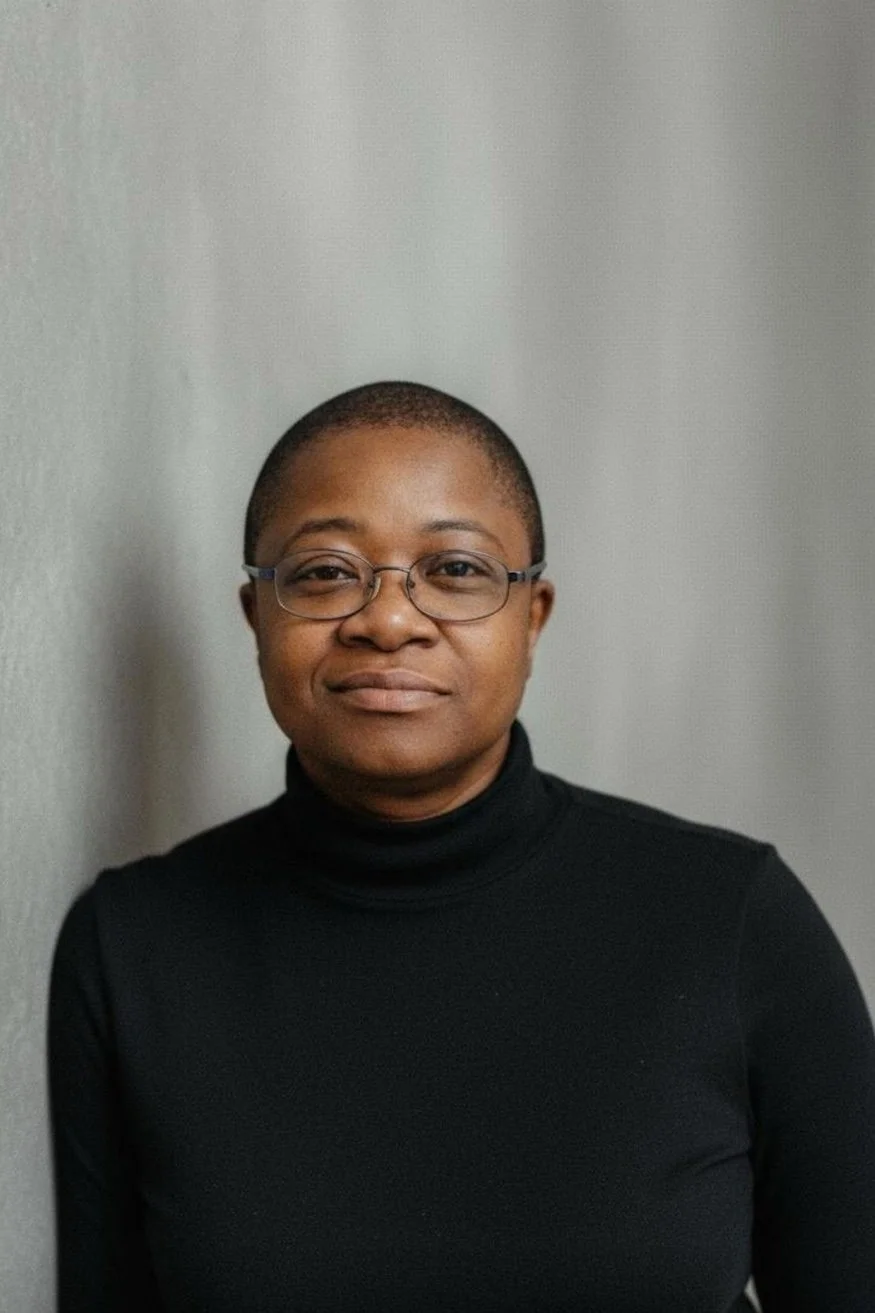 A woman with short hair, glasses, and a black turtleneck standing against a gray background.