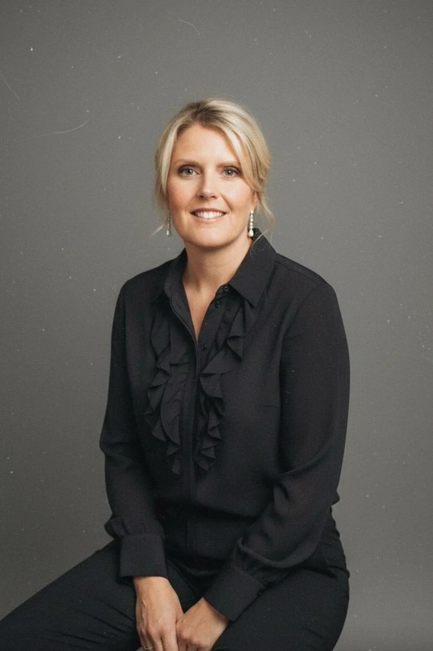 Portrait of a woman with blonde hair in a bun, wearing a black blouse with ruffles and pearl earrings, sitting in front of a gray backdrop.