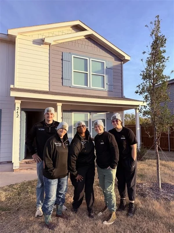 Group of five people standing in front of a two-story house with gray and beige siding and blue shutters, wearing black jackets and gray beanies, during daytime.