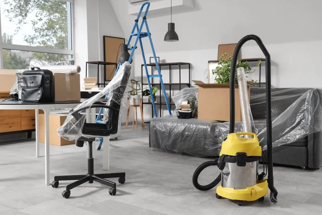 An office space being cleaned with a yellow vacuum cleaner, a black office chair, a desk with a bag, boxes, and plastic wrap, a blue ladder, and a black shelf, with large windows and plants in the background.