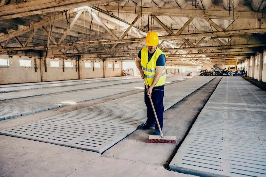 A construction worker wearing a yellow safety vest and helmet sweeps the floor of an unfinished large building with exposed wooden beams.