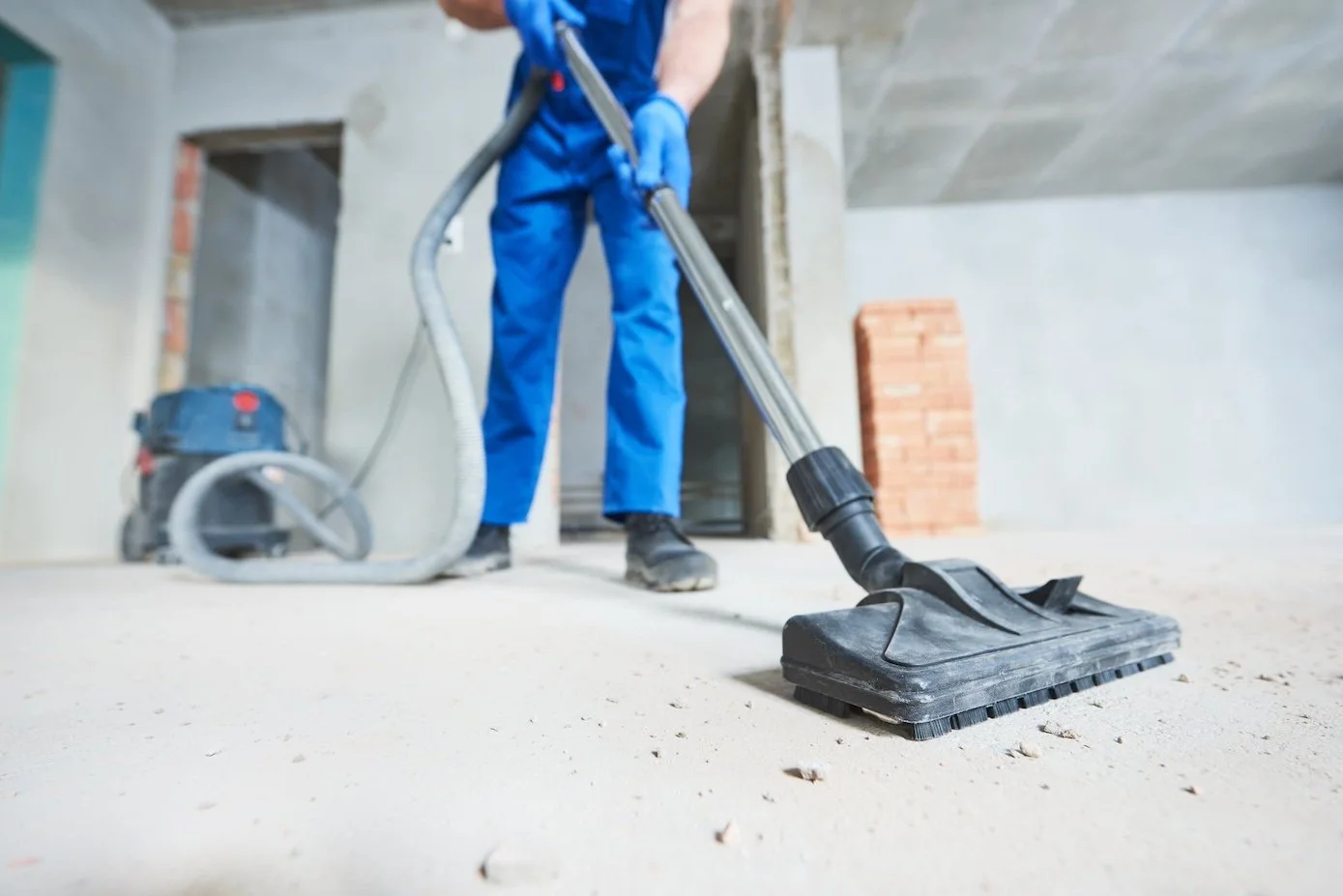 Worker in blue coveralls and gloves using a vacuum to clean an unfinished concrete floor in a building under construction.