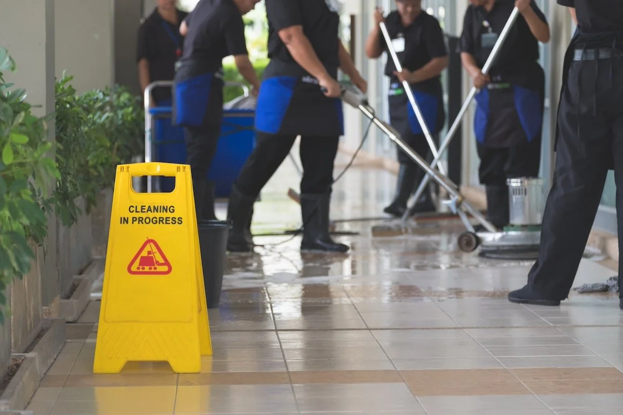 Cleaning crew working on tiled floor with a yellow 'Cleaning in Progress' sign in the foreground.