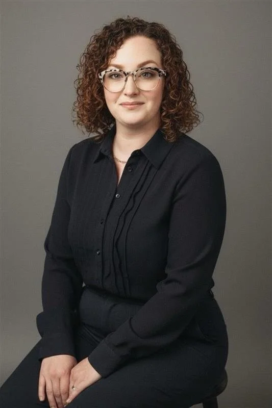 A woman with curly brown hair and glasses, wearing a black long-sleeve shirt, sitting against a plain gray background.