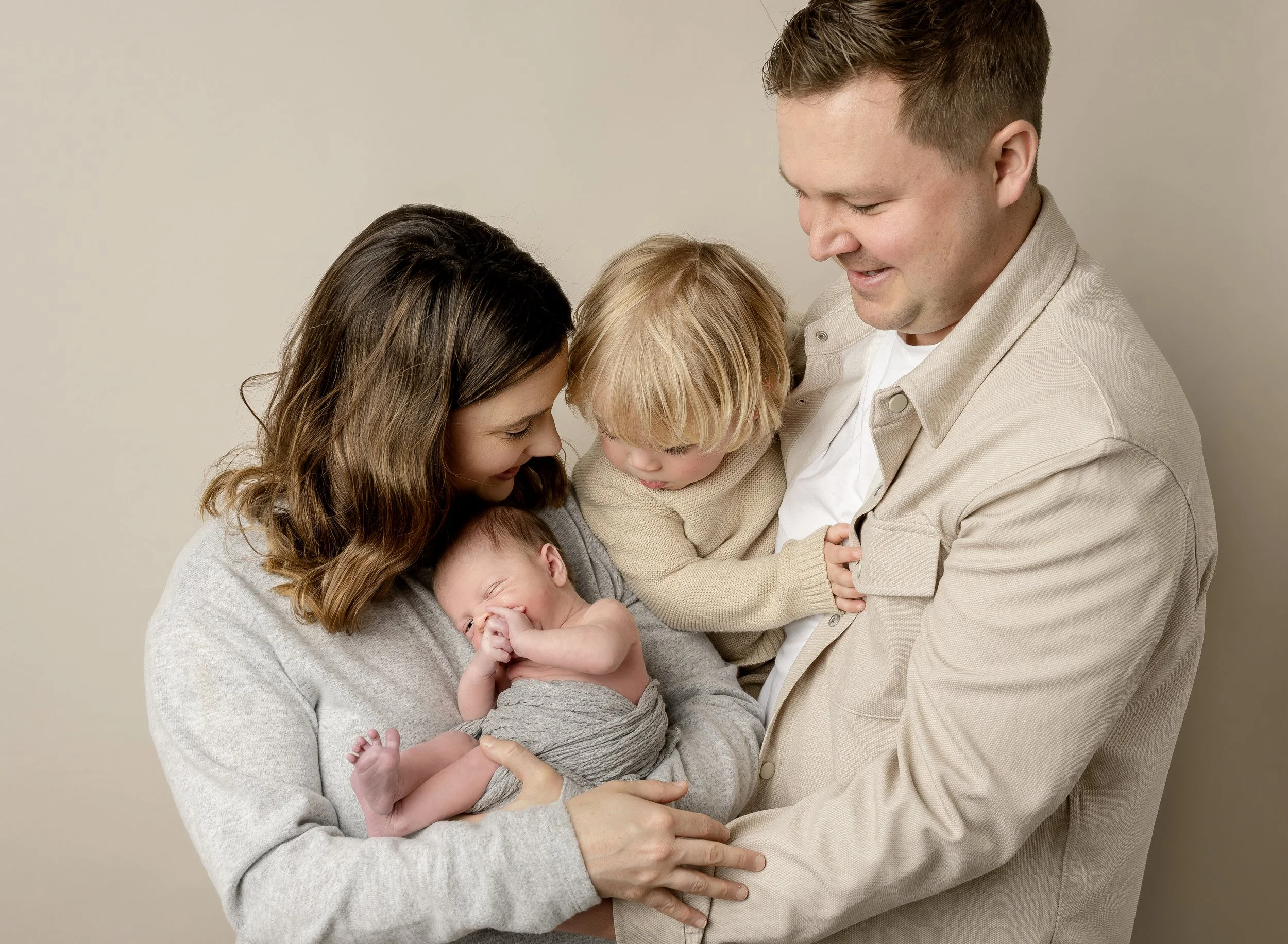 A happy family of four, including a mother with brown hair, a father with short hair, and two young children, a toddler and a newborn, together in a close embrace against a neutral background.