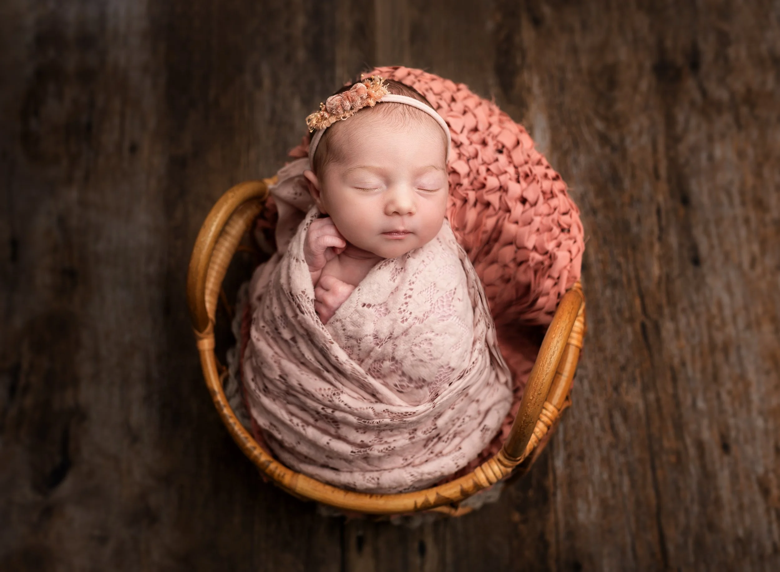 A sleeping newborn baby wrapped in a pink lace blanket, wearing a floral headband, in a wicker basket on a wooden floor.