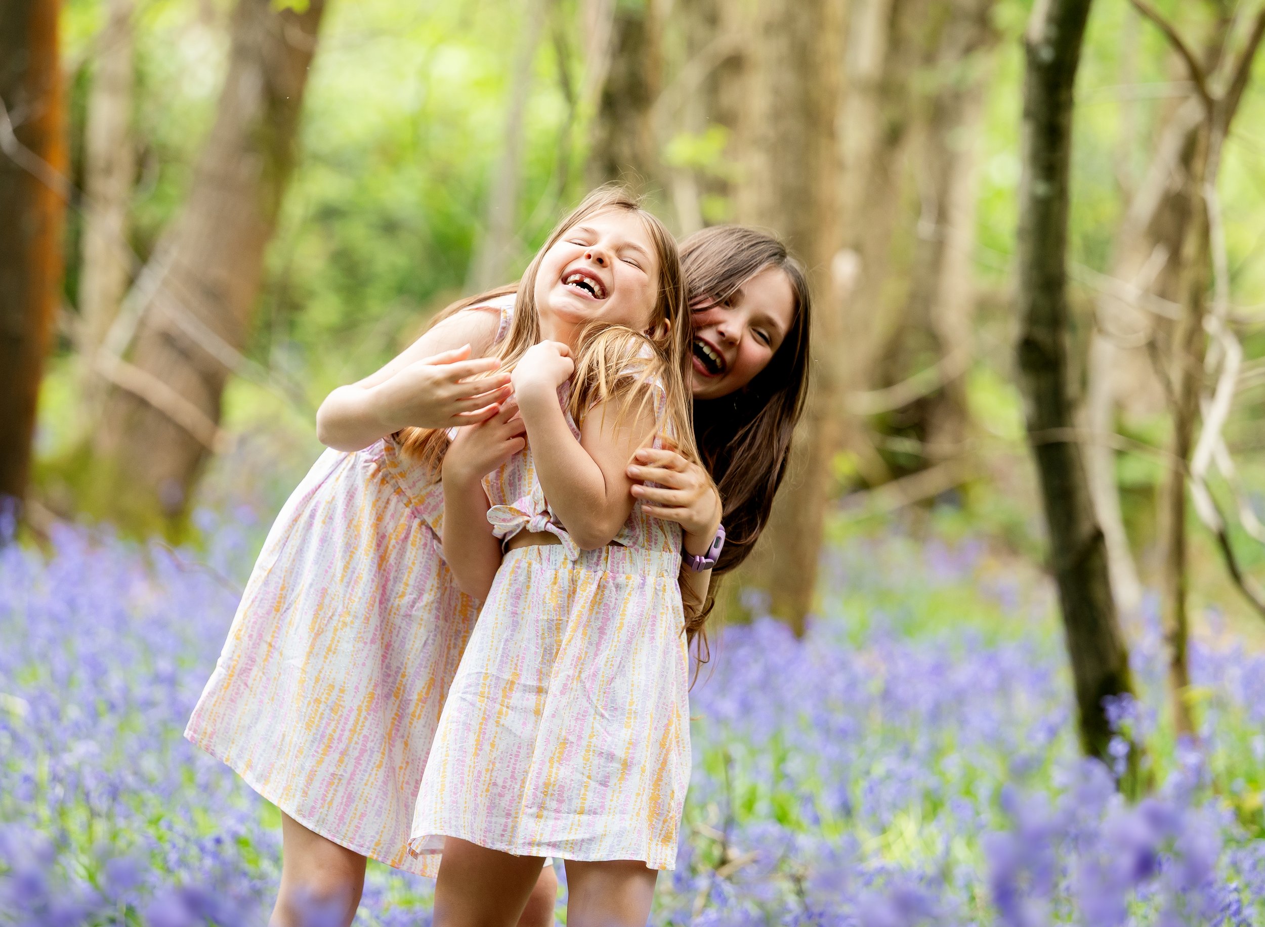 Two girls laughing and hugging in a forest with trees and bluebell flowers.