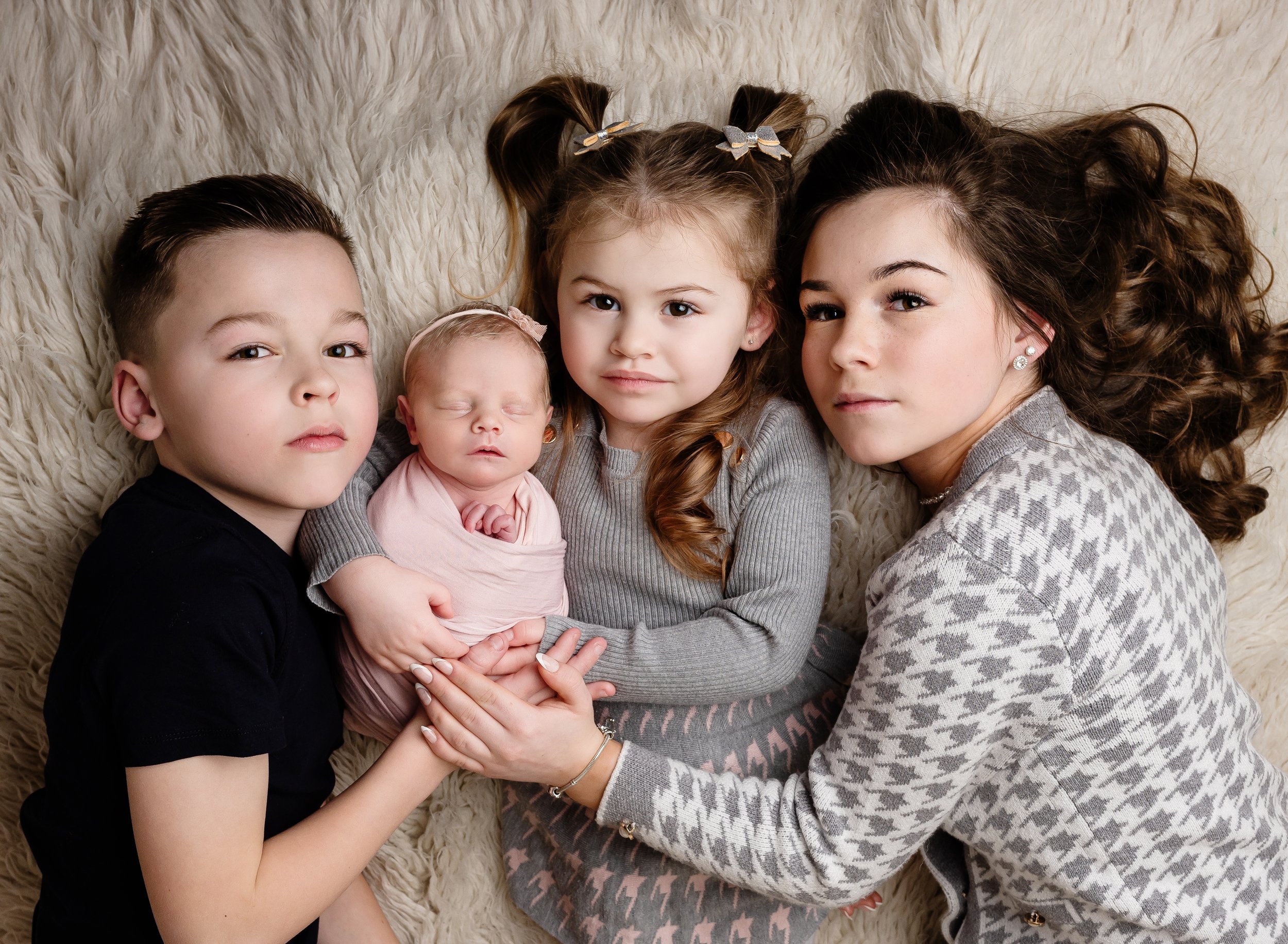 Four children lying on a fluffy cream-colored rug, with a newborn baby in the center. The baby is wrapped in pink cloth, sleeping with eyes closed. An older girl with brown hair in pigtails and bows is holding the baby, and an older boy with short ha