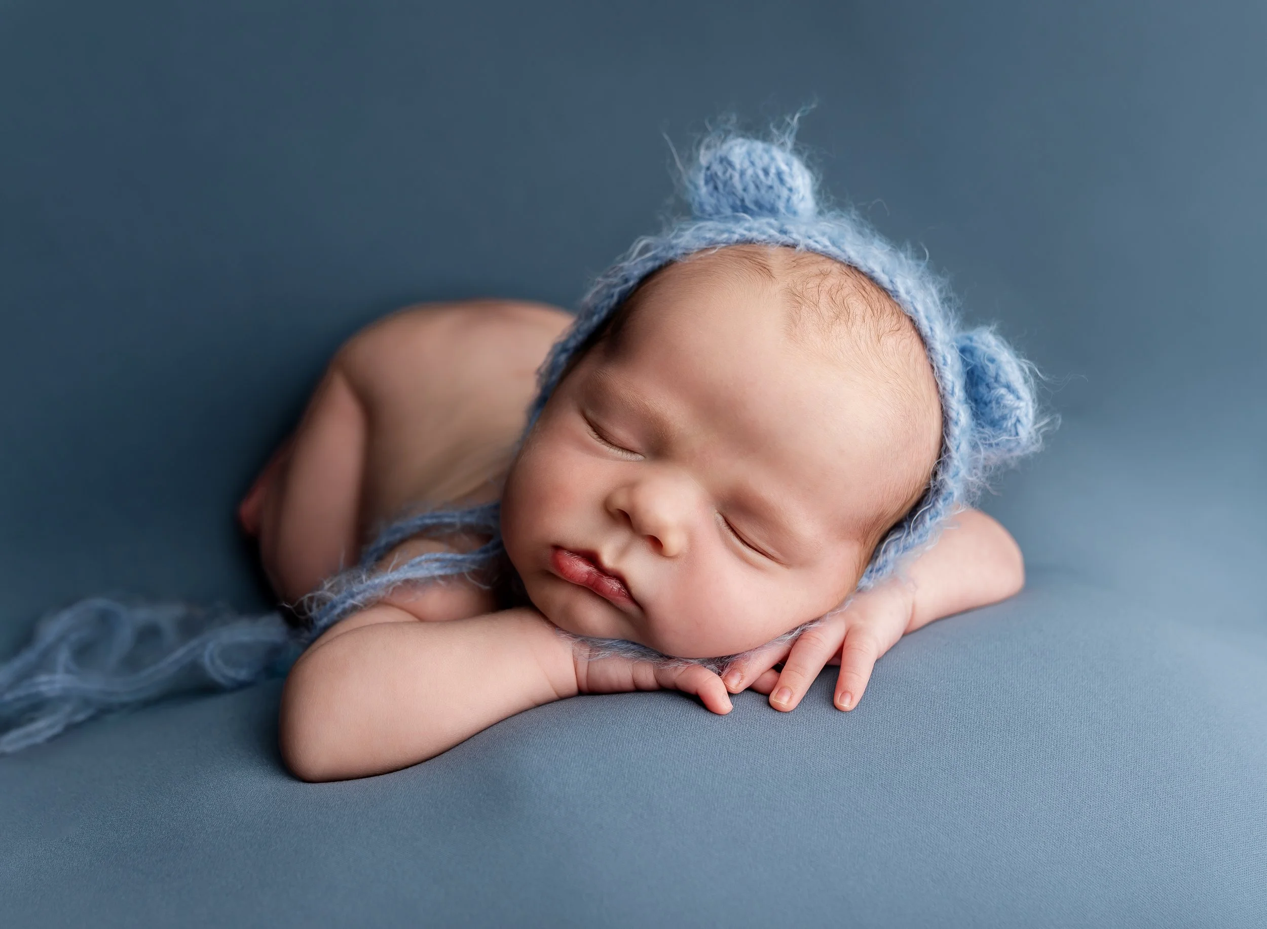 A sleeping baby with a light blue knit hat with bear ears, lying on a soft blue surface.