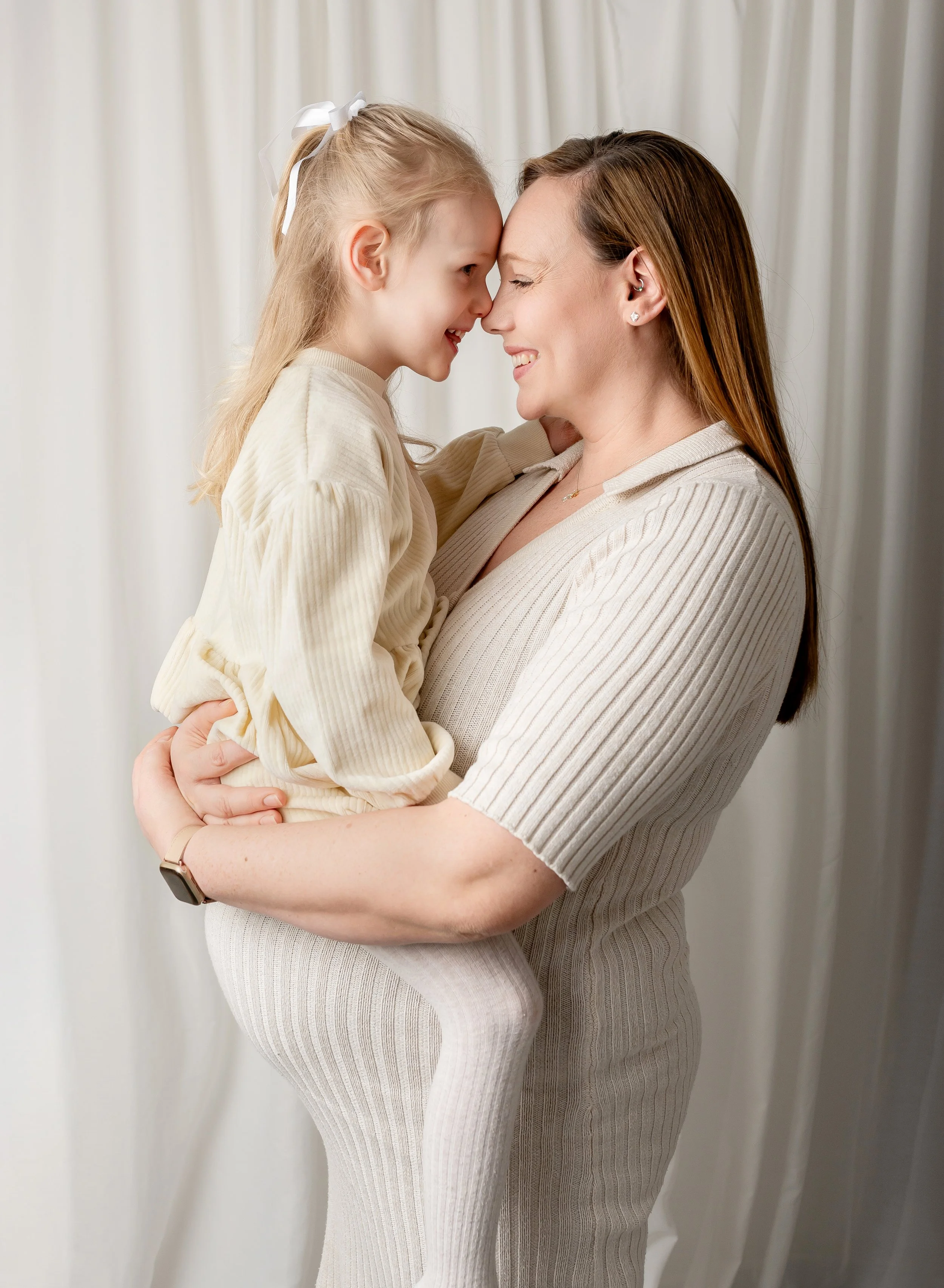 A woman and girl smiling while touching foreheads, with the woman holding the girl up in front of a white curtain.