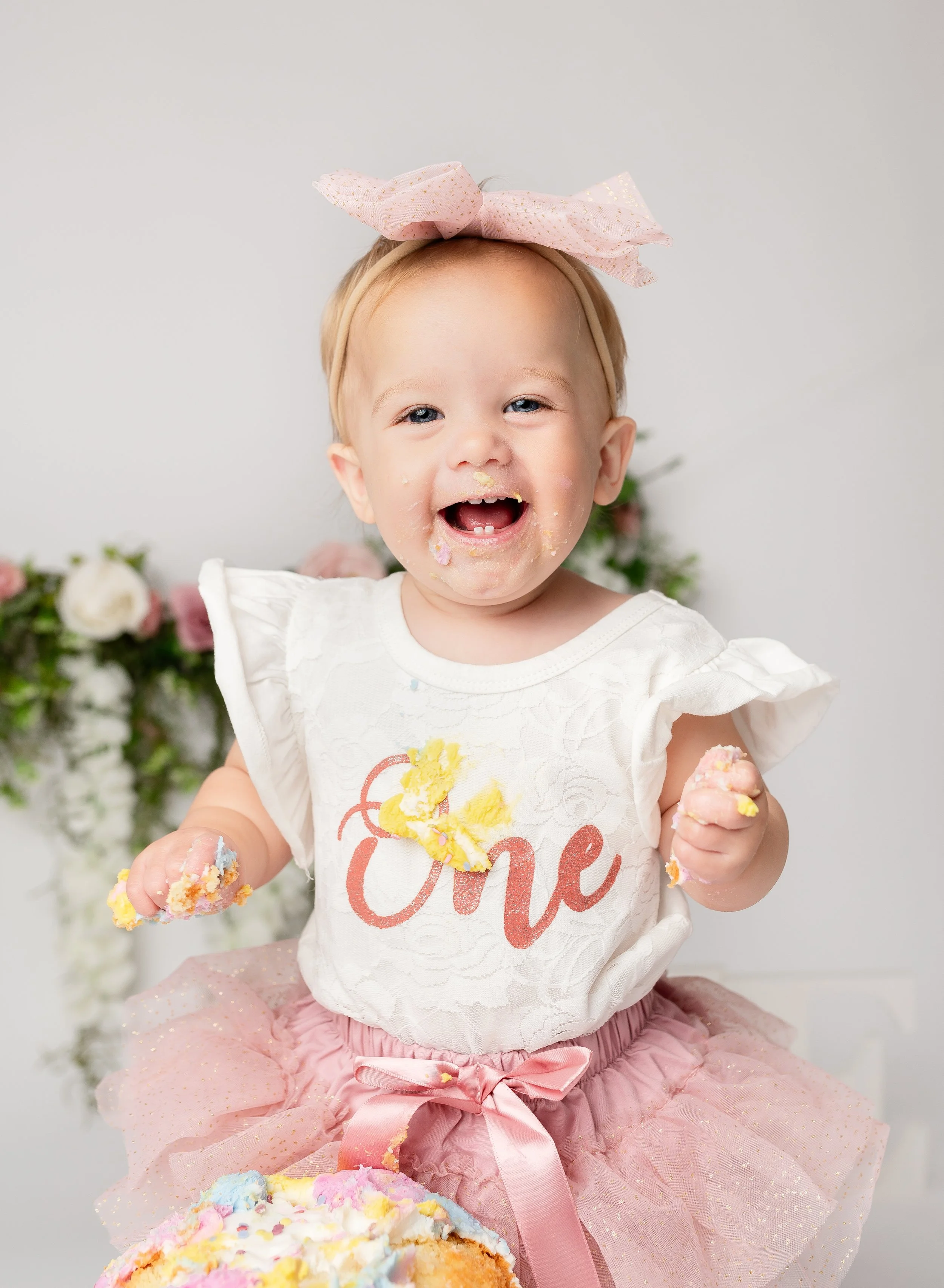 A young girl wearing a white shirt with the word 'One' and a pink tutu, celebrating her first birthday with cake and frosting on her face and hands, smiling joyfully.
