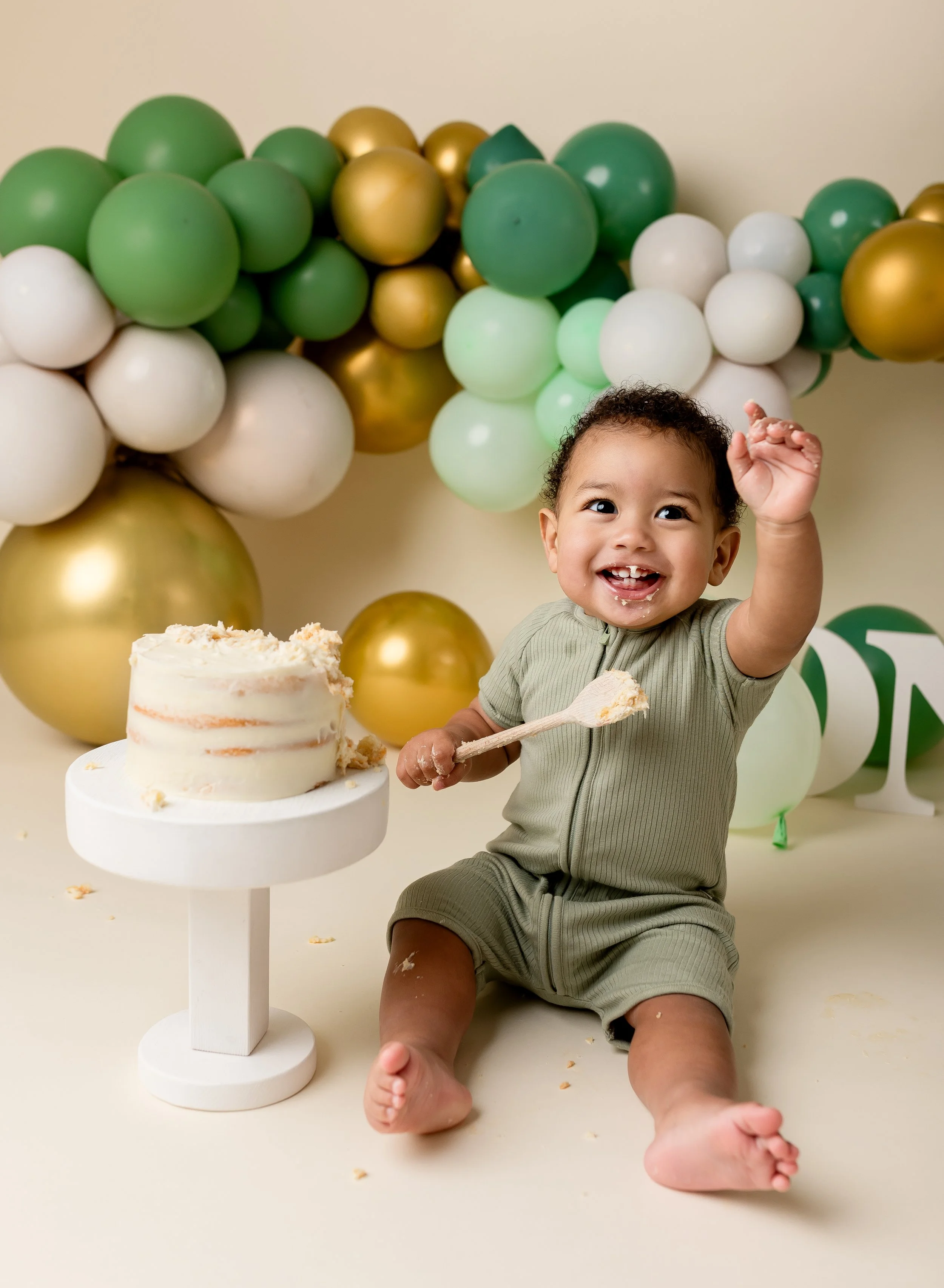 A smiling toddler seated on the floor with a small birthday cake on a white pedestal, surrounded by green, white, and gold balloons, in a celebration setting.