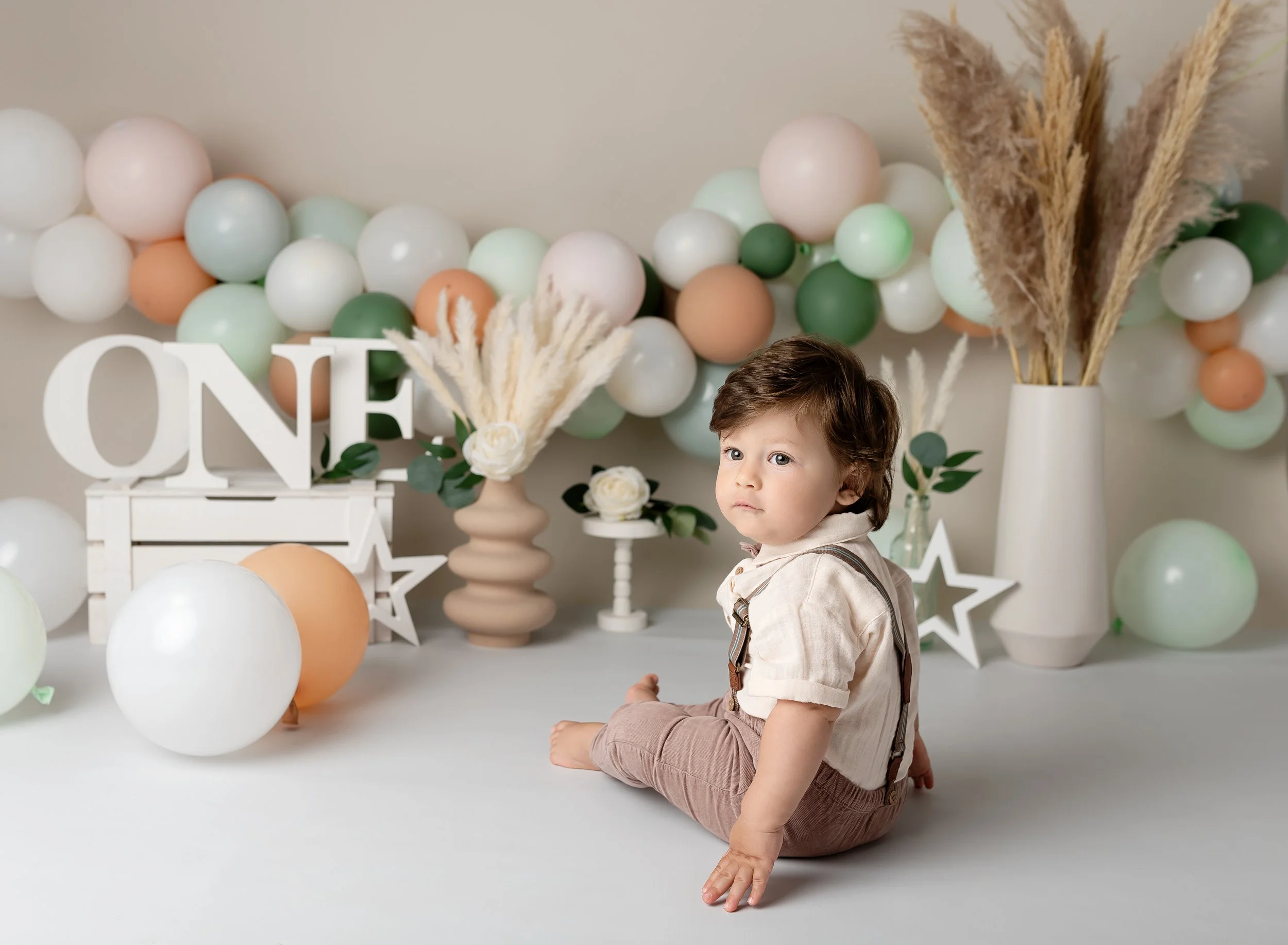 Young child sitting on the floor in front of pastel birthday decorations, including balloons, flowers, and a "ONE" sign, with neutral and soft colors.