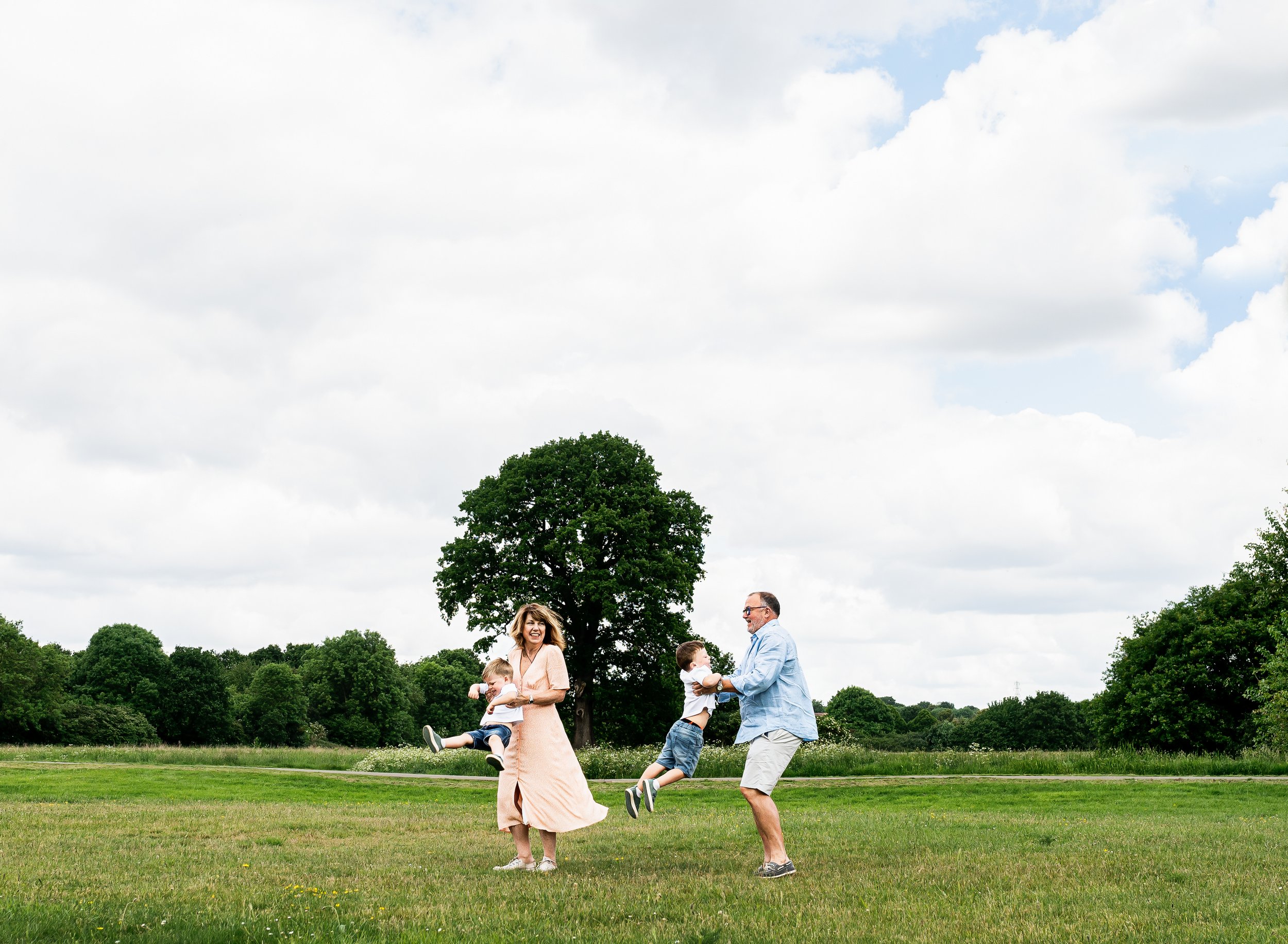 Family of four playing and laughing on a grassy field outdoors beneath a partly cloudy sky with trees in the background.