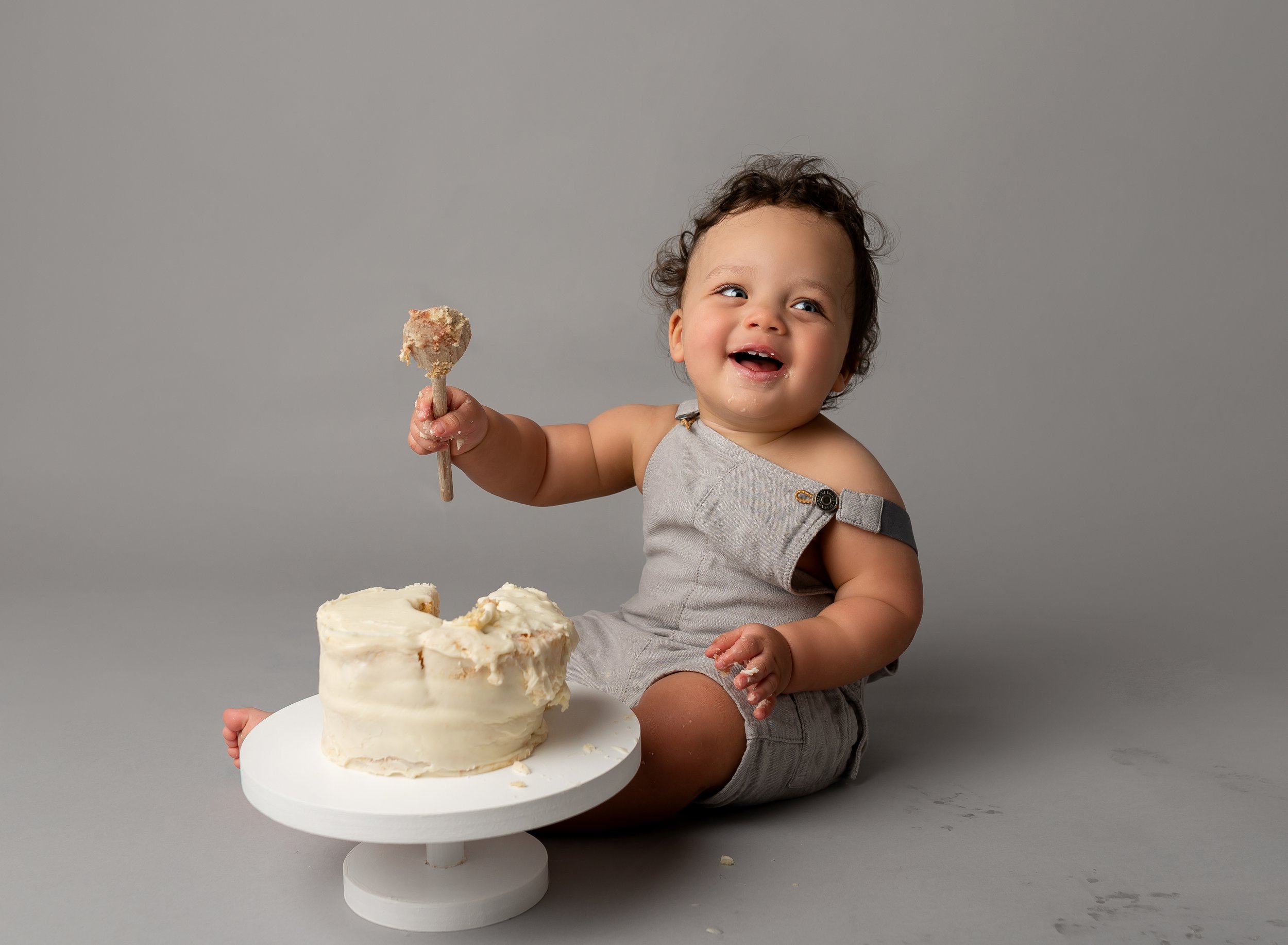 Smiling toddler in gray outfit sitting on gray background, holding a cake knife with cake crumbs, next to a partially eaten white frosted cake on a white stand.