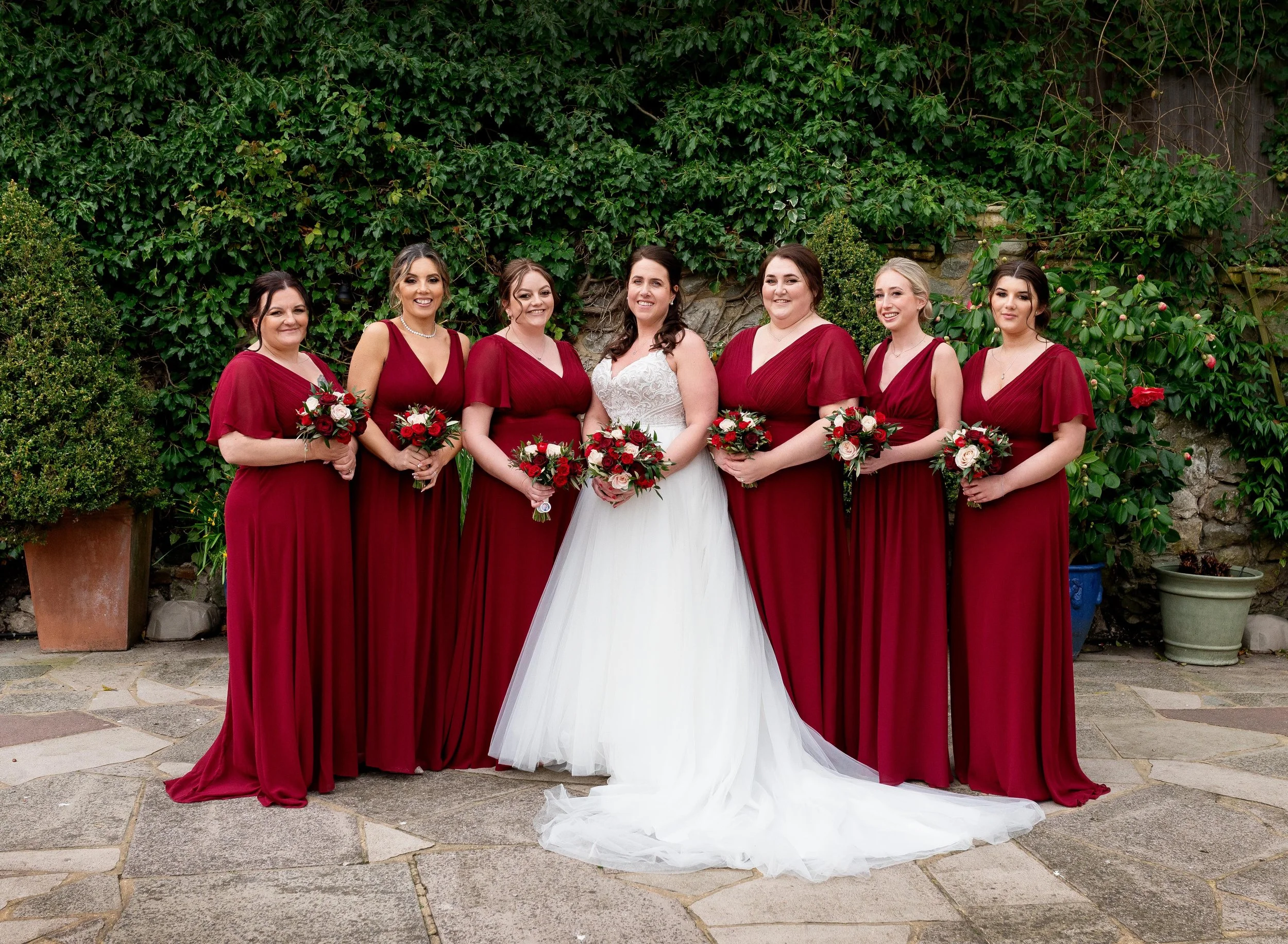 Bride in a white gown with six bridesmaids in matching red dresses, holding bouquets, standing outdoors against a green leafy background.