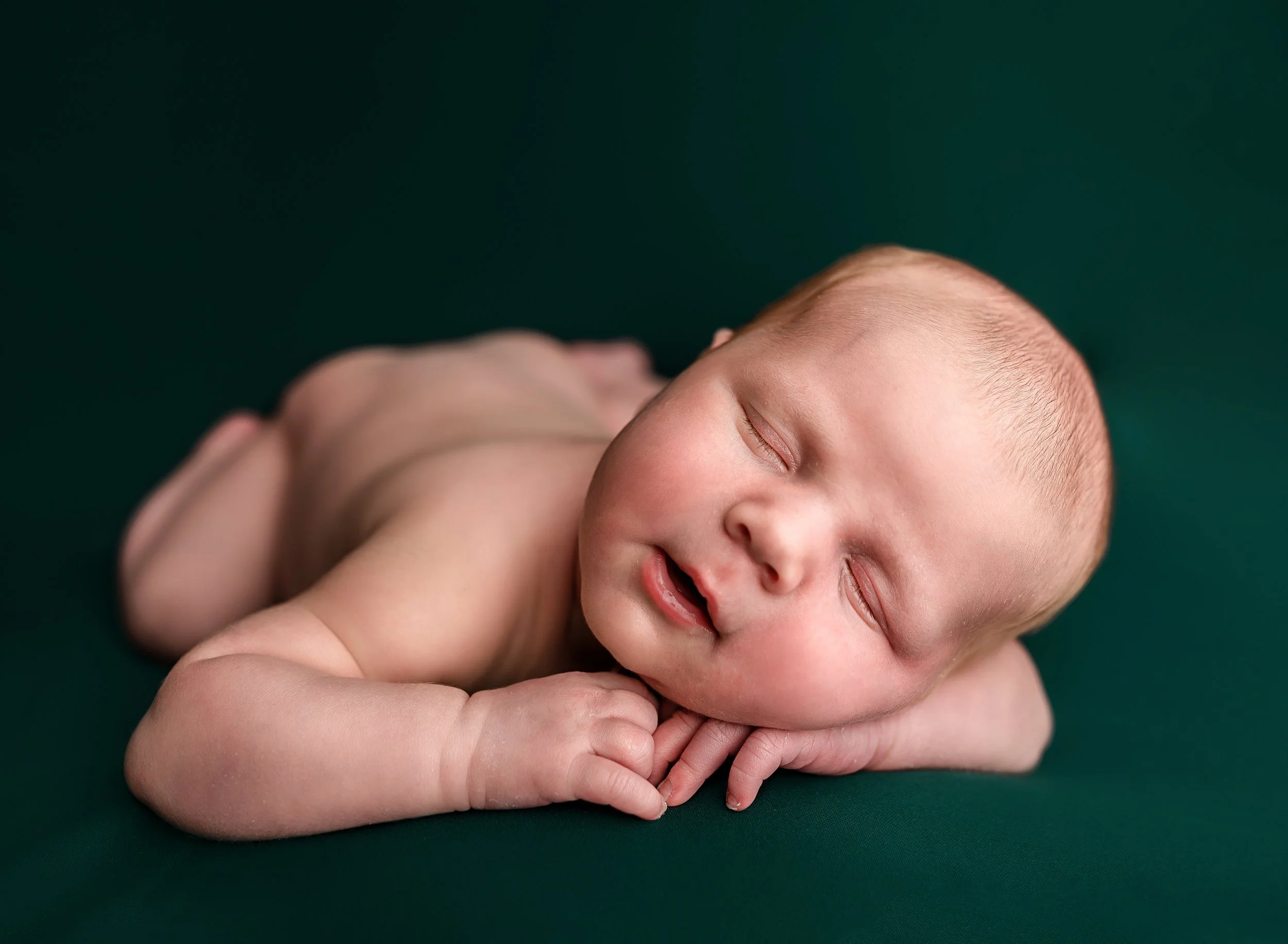 A newborn baby sleeps peacefully on a dark green surface with hands tucked under its cheek.