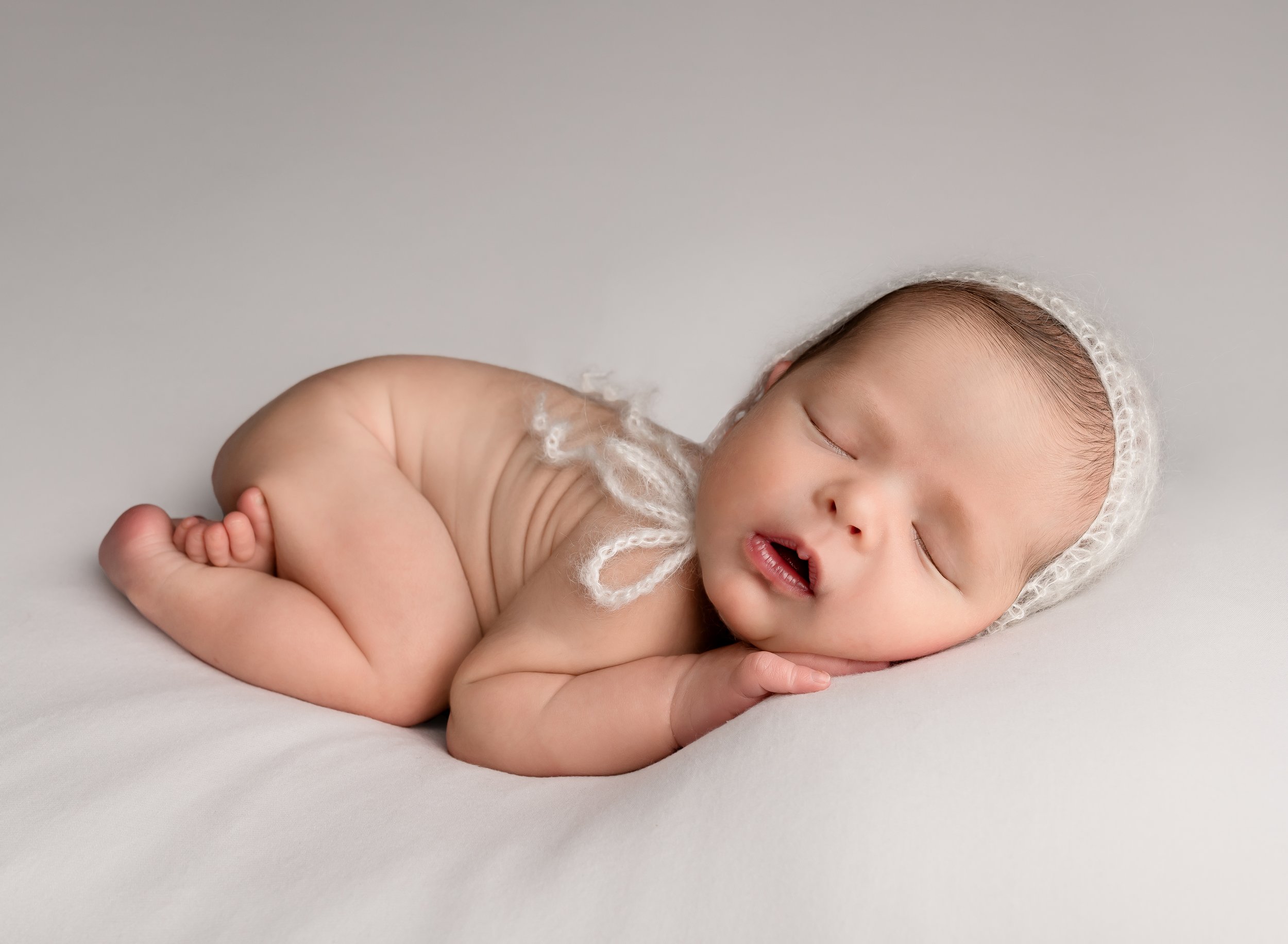 A sleeping newborn baby girl lying on her side on a soft white surface, wearing a white knitted hat, with her hands tucked under her face and her legs curled up.