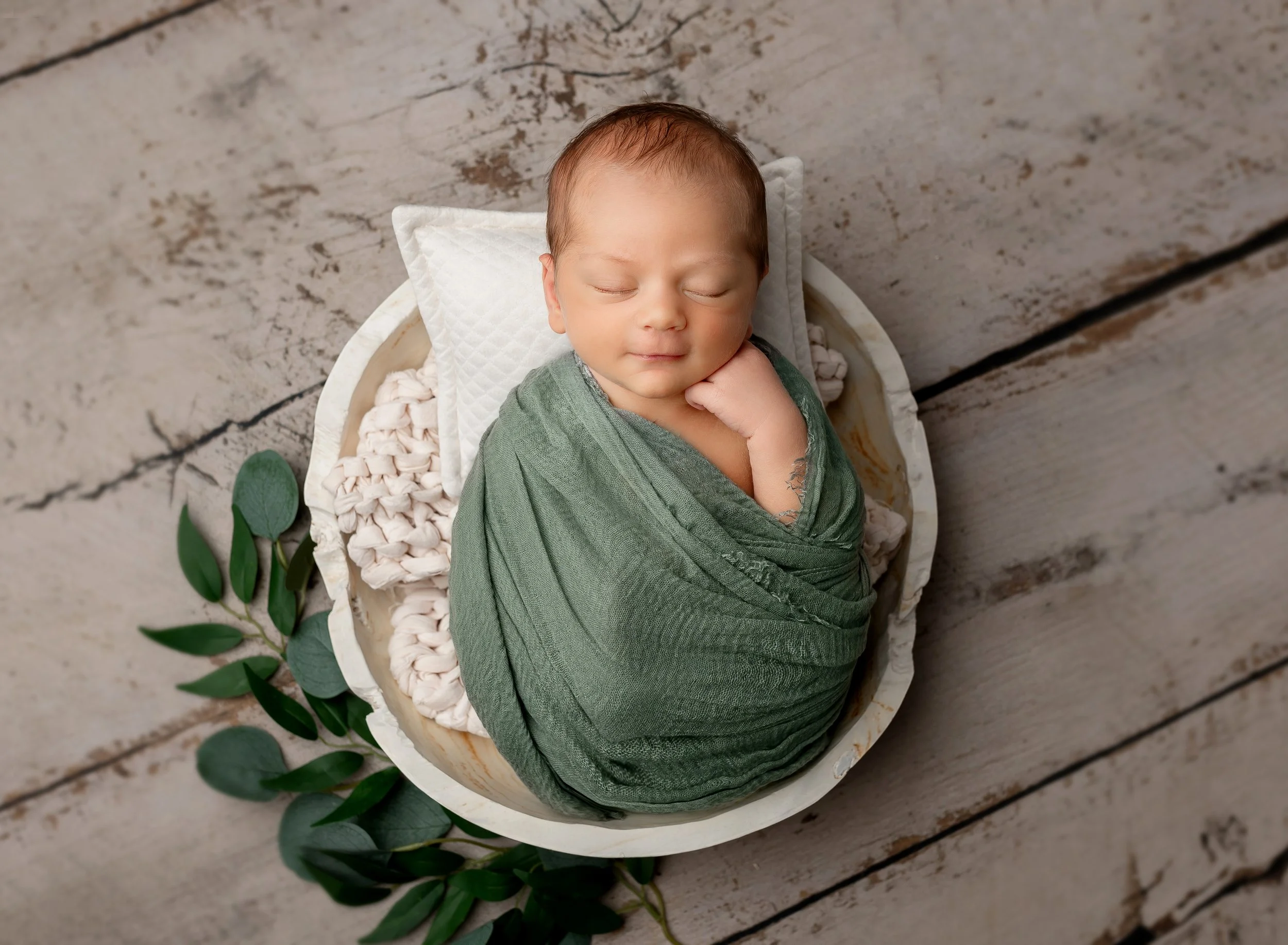 A newborn baby wrapped in a green blanket, lying on a white quilted pillow inside a round basket, on a rustic wooden floor, with green leaves beside the basket.