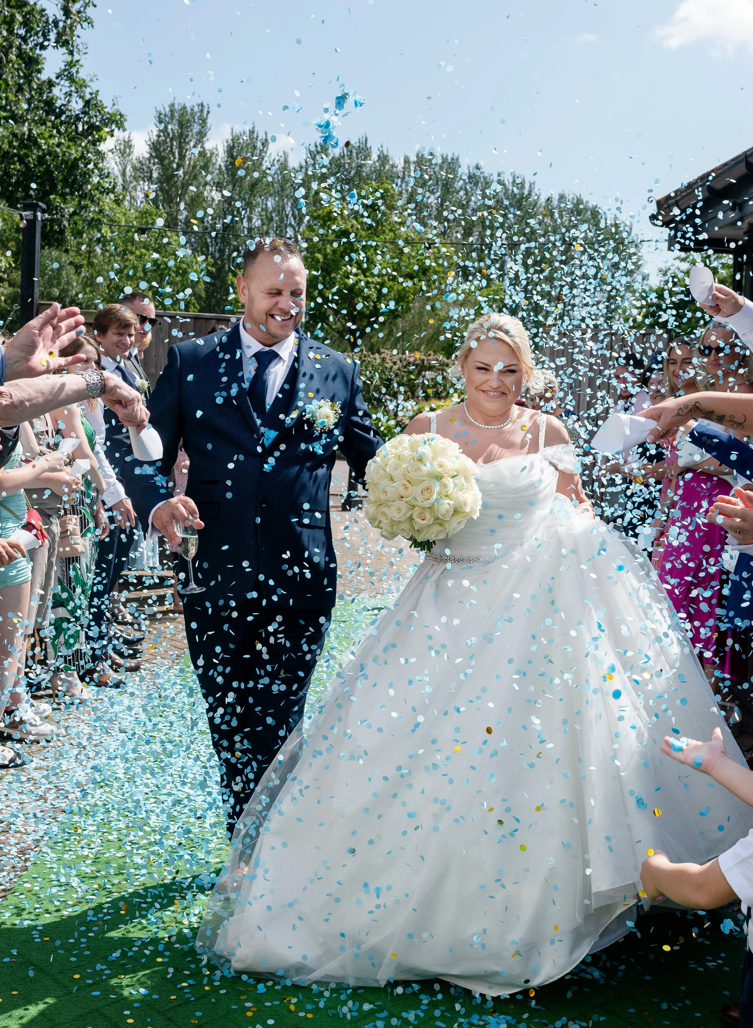 A newlywed couple walking through a shower of blue and gold confetti, surrounded by friends and family, on a sunny outdoor wedding day. The bride holds a bouquet of white roses and wears a white wedding gown, and the groom wears a navy blue suit. Eve