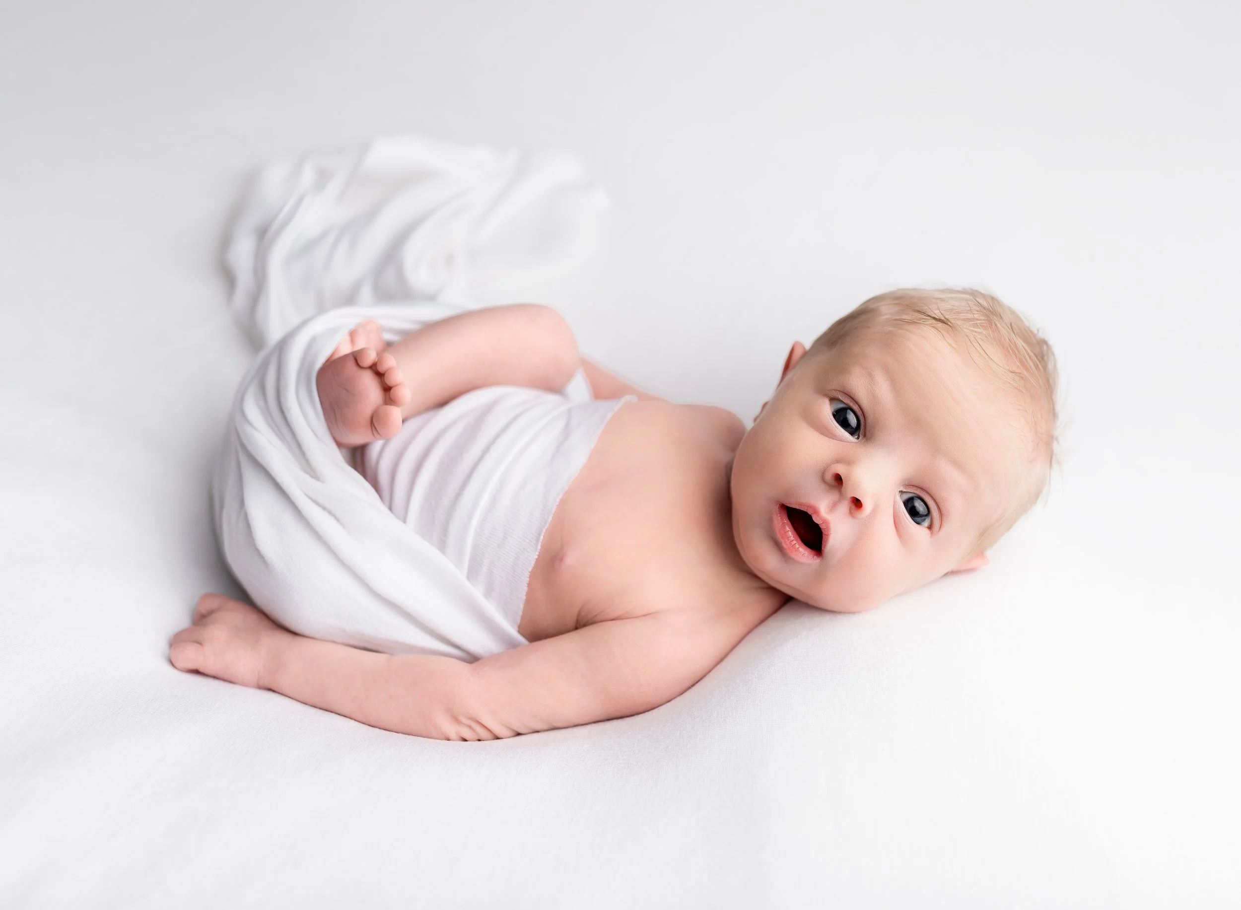 A baby lying on a white surface, with a white cloth draped over their lower body, looking at the camera with an open mouth and wide eyes.