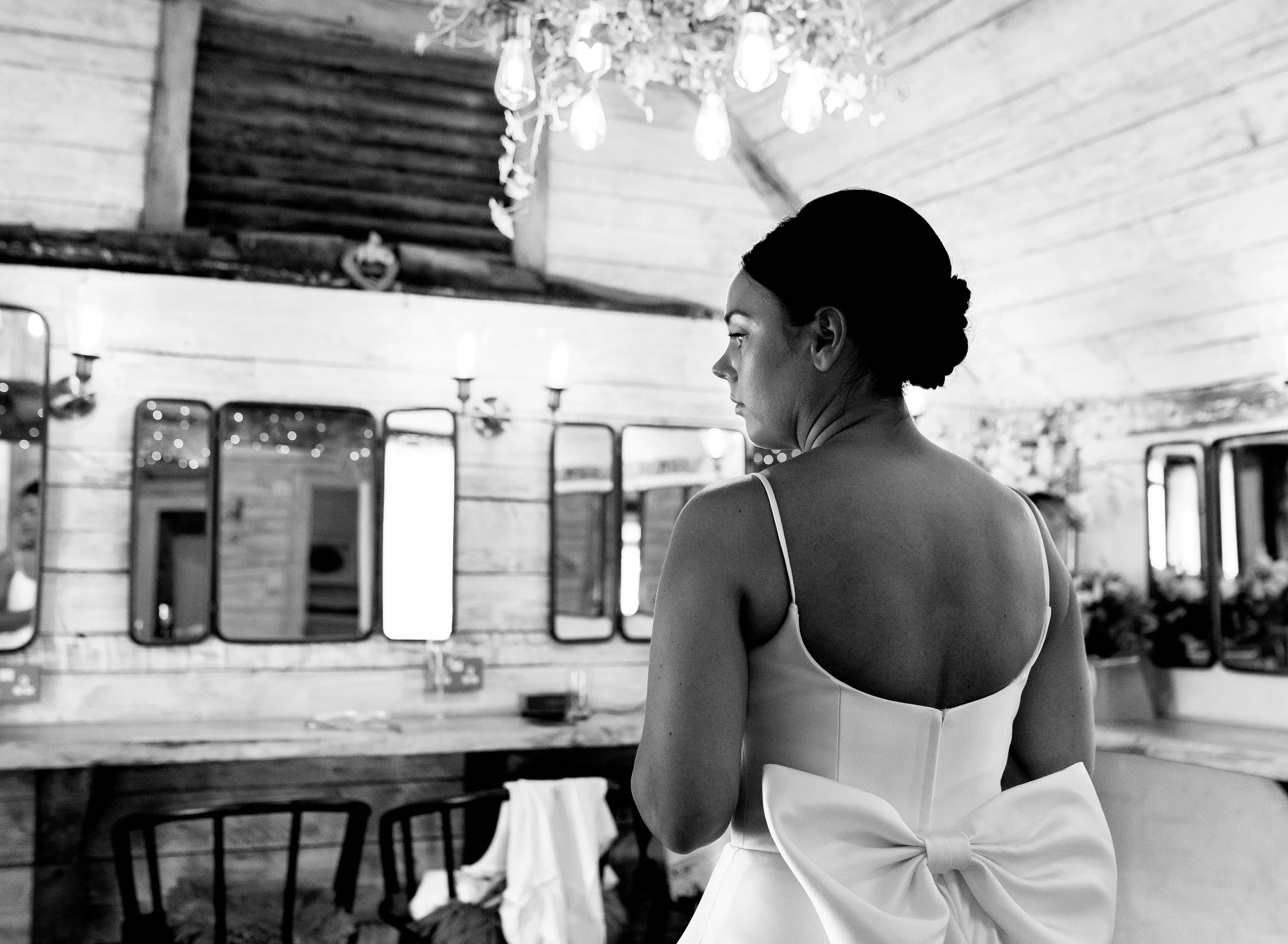 A woman in a white dress with a large bow on the back standing in a rustic room with mirror, candles, and string lights.